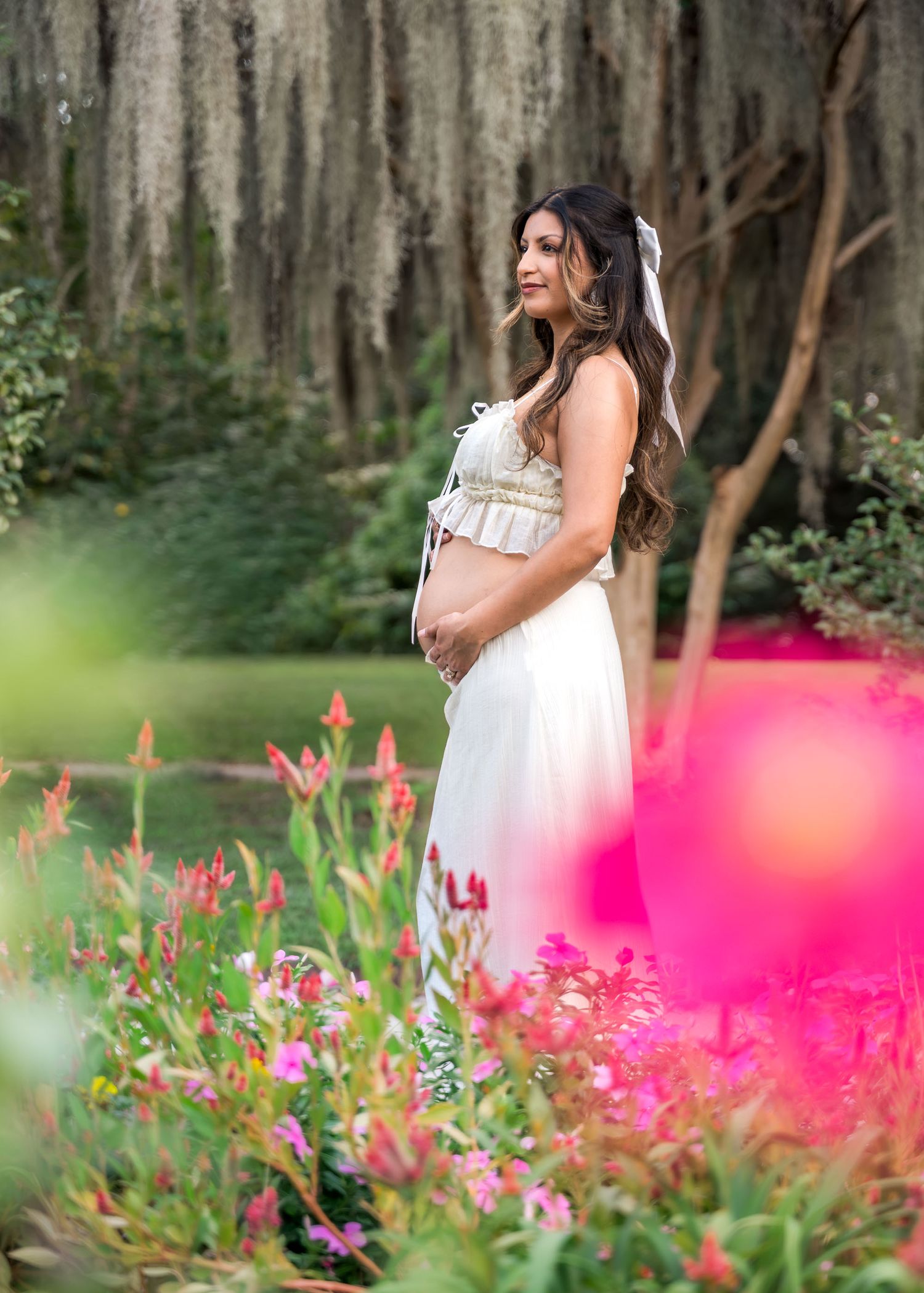 Hampton Park. Elegant pregnant woman in white gown posing among vibrant pink flowers in garden.