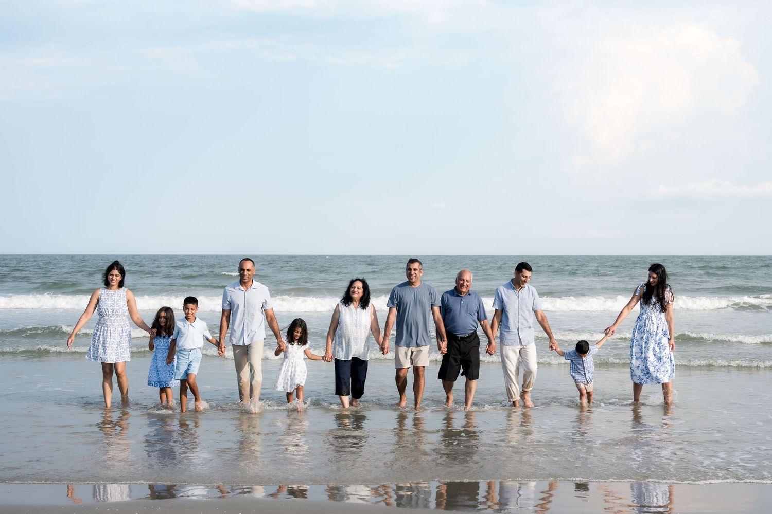 Large family holding hands on a sandy beach with ocean waves, wearing coordinated blue and white outfits.