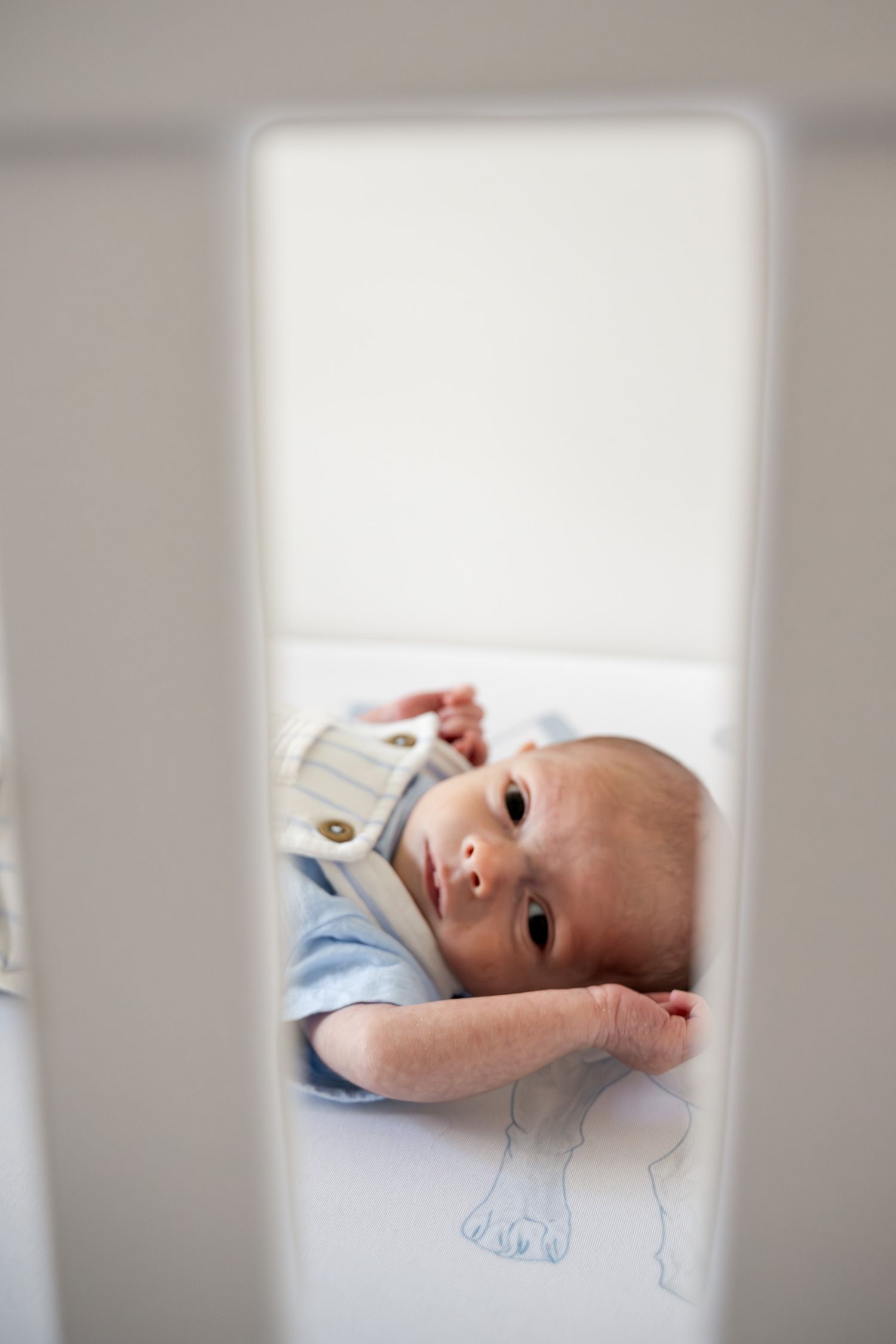 Curious newborn baby in blue outfit peeking through white crib slats with wide eyes.