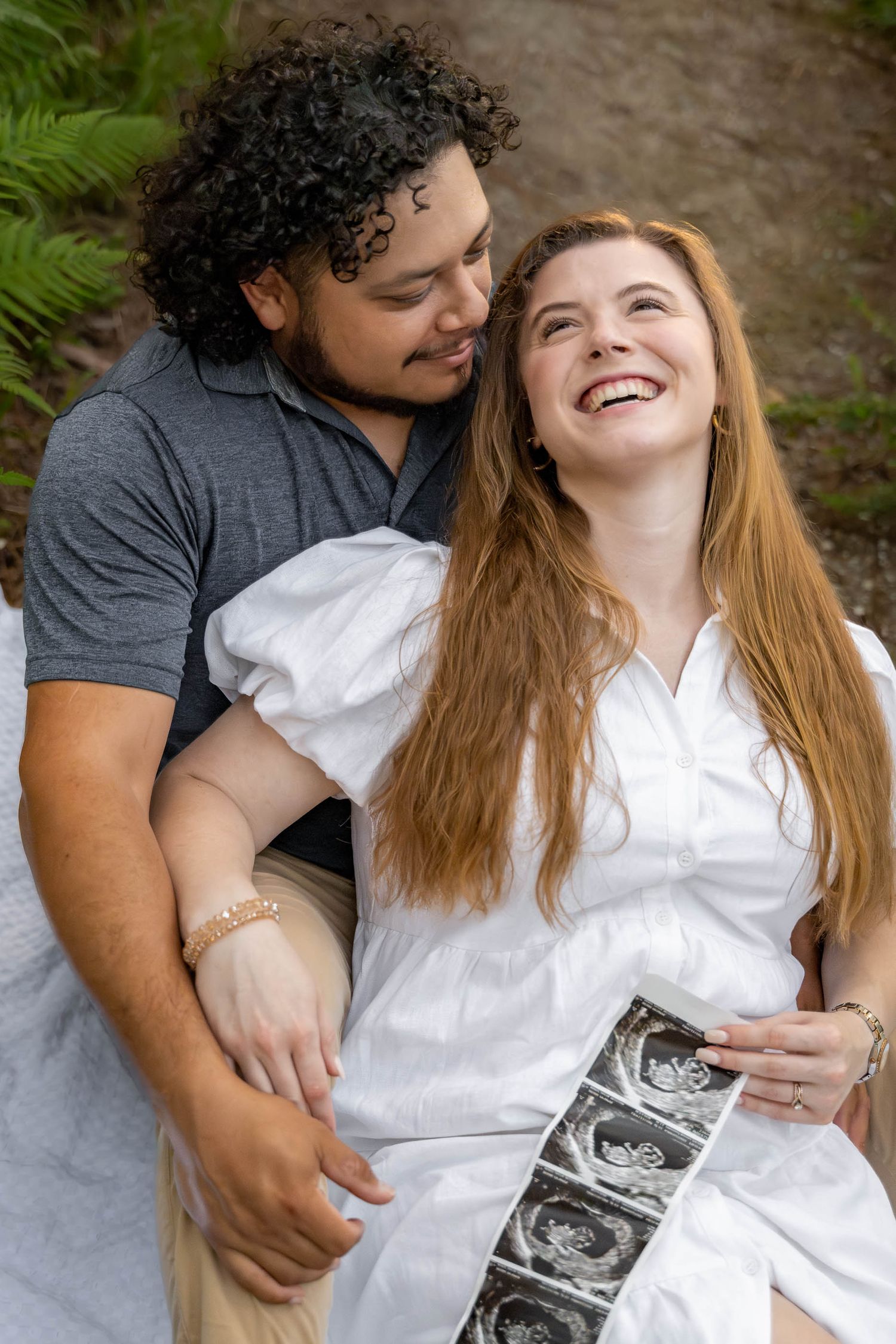 Joyful couple holding ultrasound photos during maternity shoot in lush green forest setting.