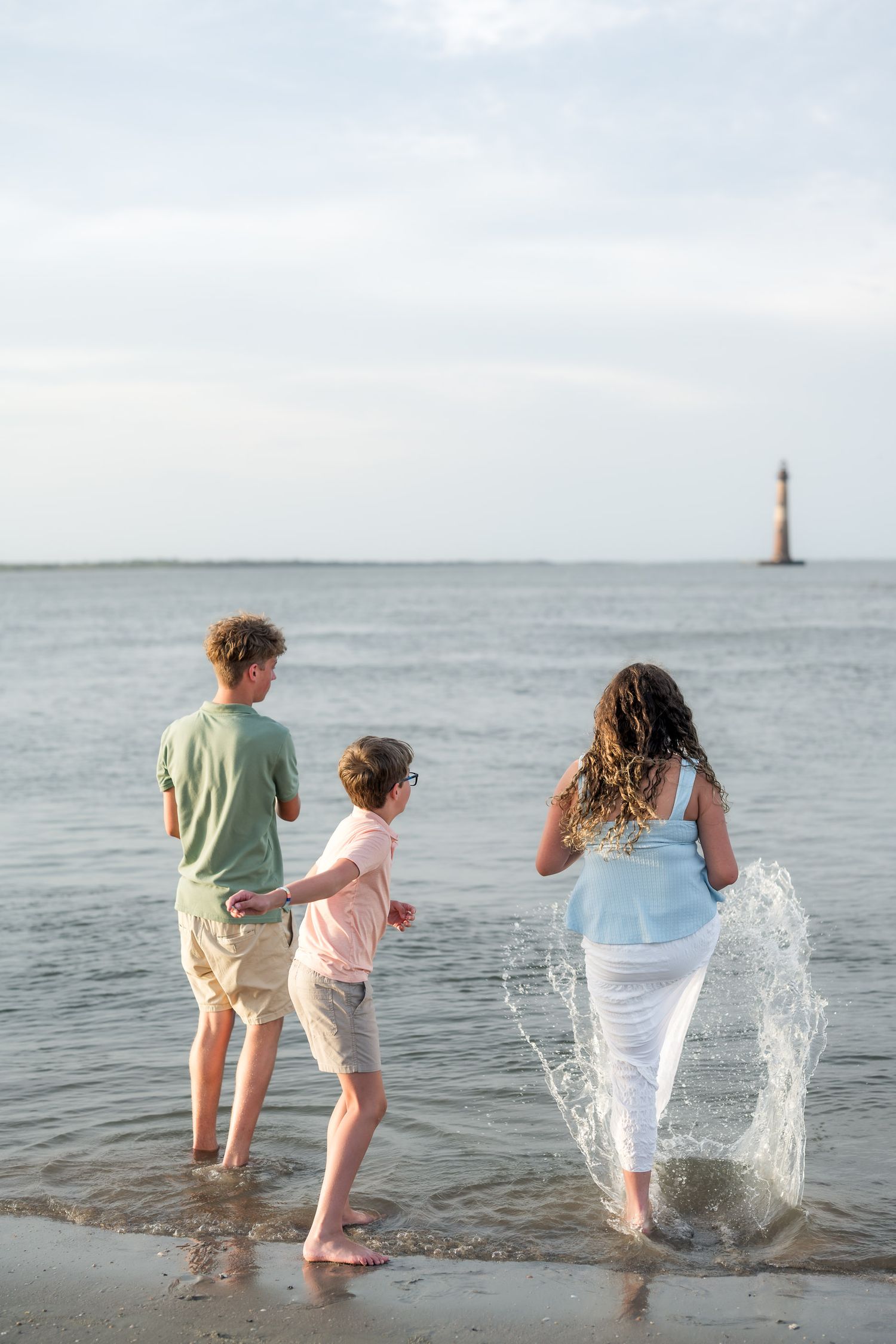 Three children playing in ocean waves on a beach with a distant lighthouse on a calm day.
