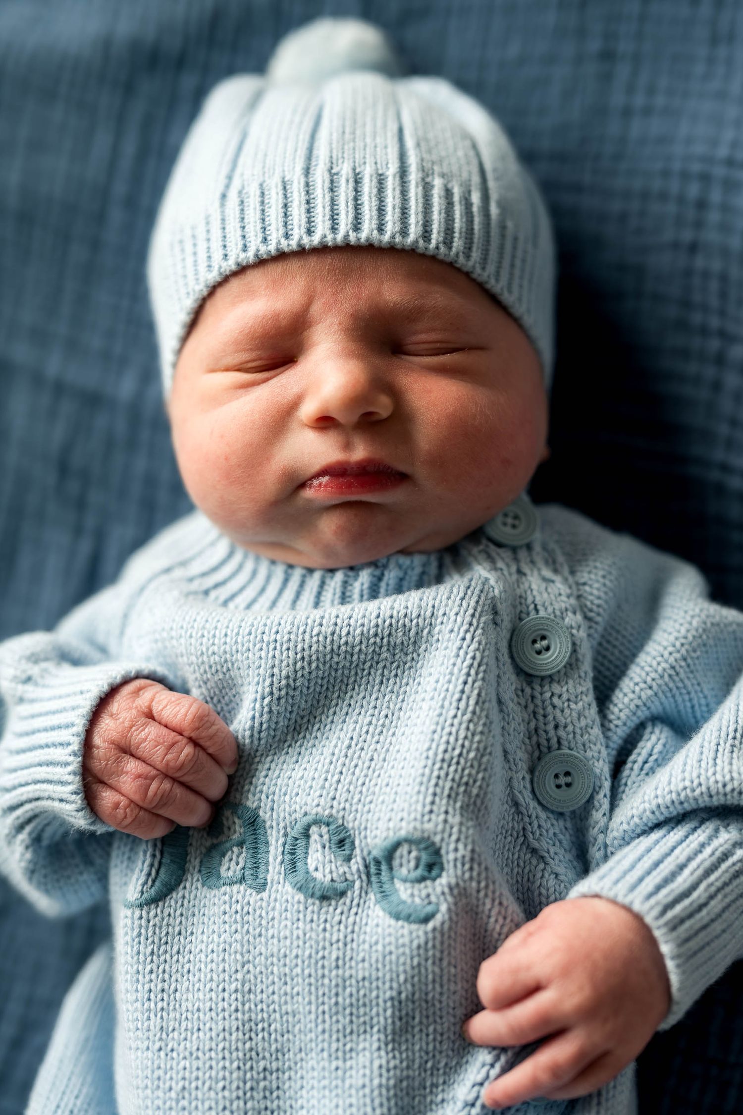 Sleeping newborn baby wearing a light blue knit sweater and matching hat with a pompom on top.