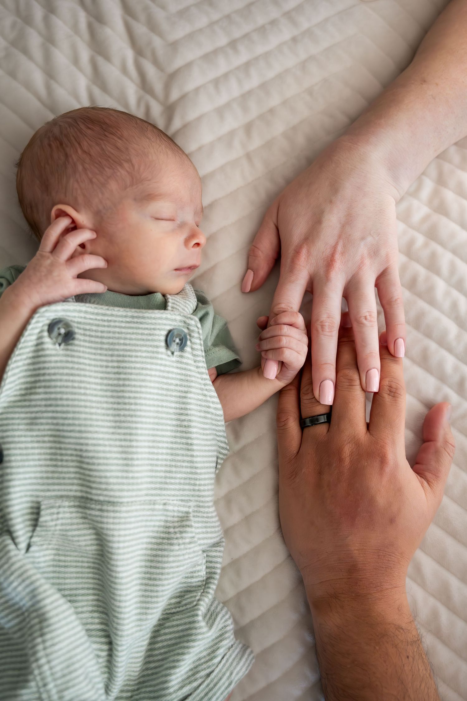 Sleeping newborn in green onesie holds parents hands on a white quilted blanket.