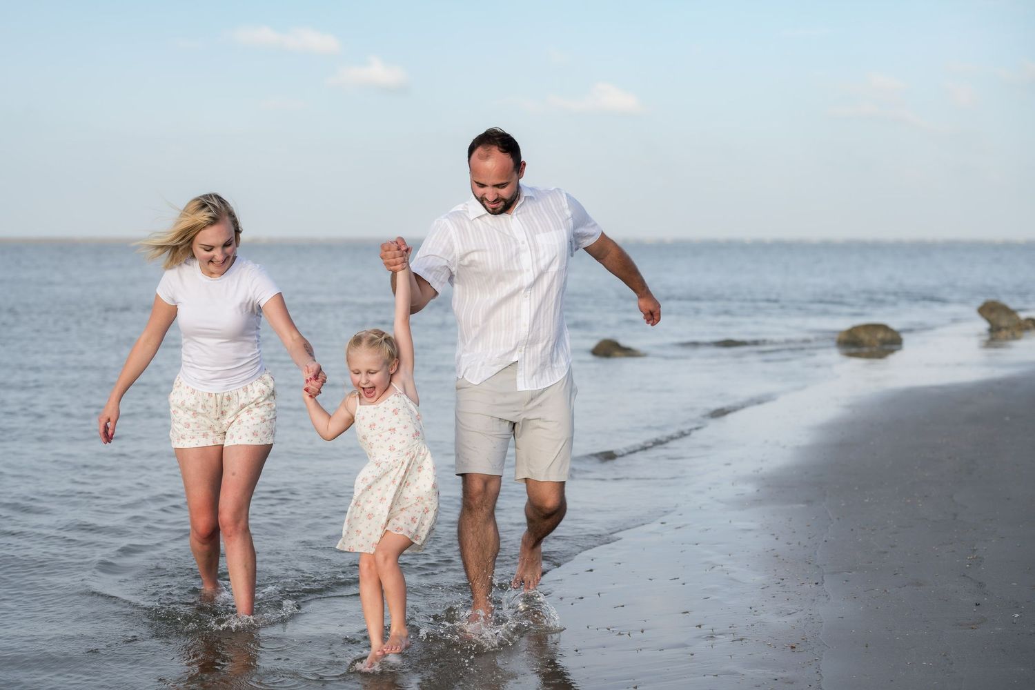 Happy family of three splashing in ocean waves on a sunny beach, parents holding young daughter's hands.