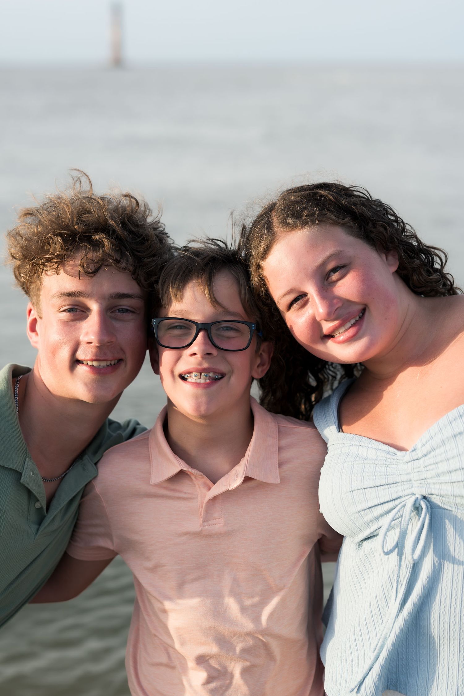Three smiling kids pose together on a beach, middle child wearing glasses and braces, ocean visible behind them.