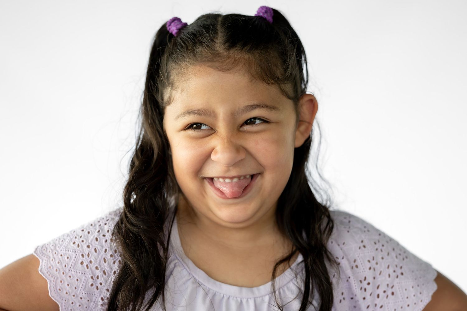 Smiling young Latina girl with dark hair in pigtails wearing a white top against a light background.