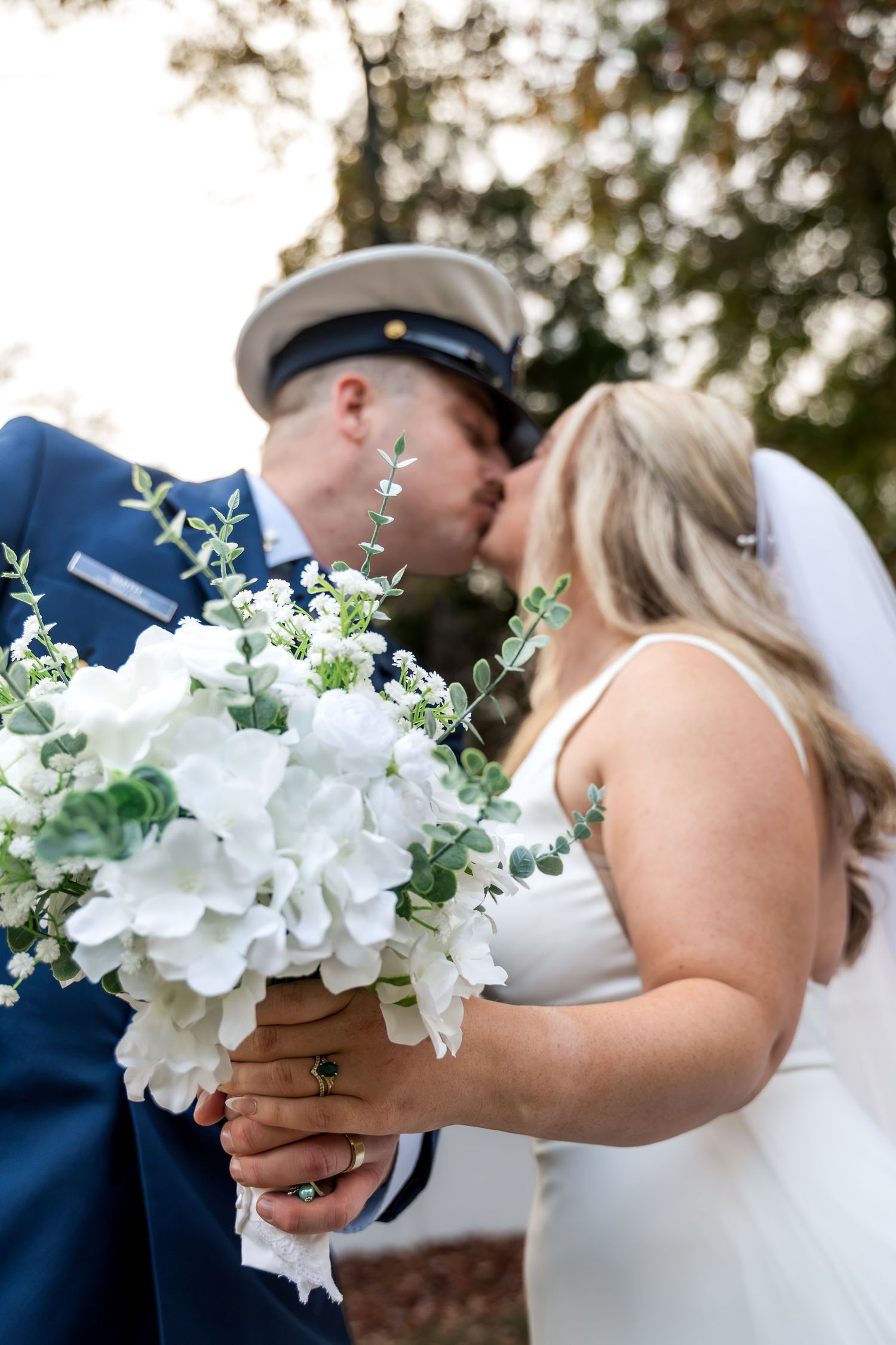 Newlyweds sharing first kiss behind white hydrangea bridal bouquet at outdoor wedding ceremony.
