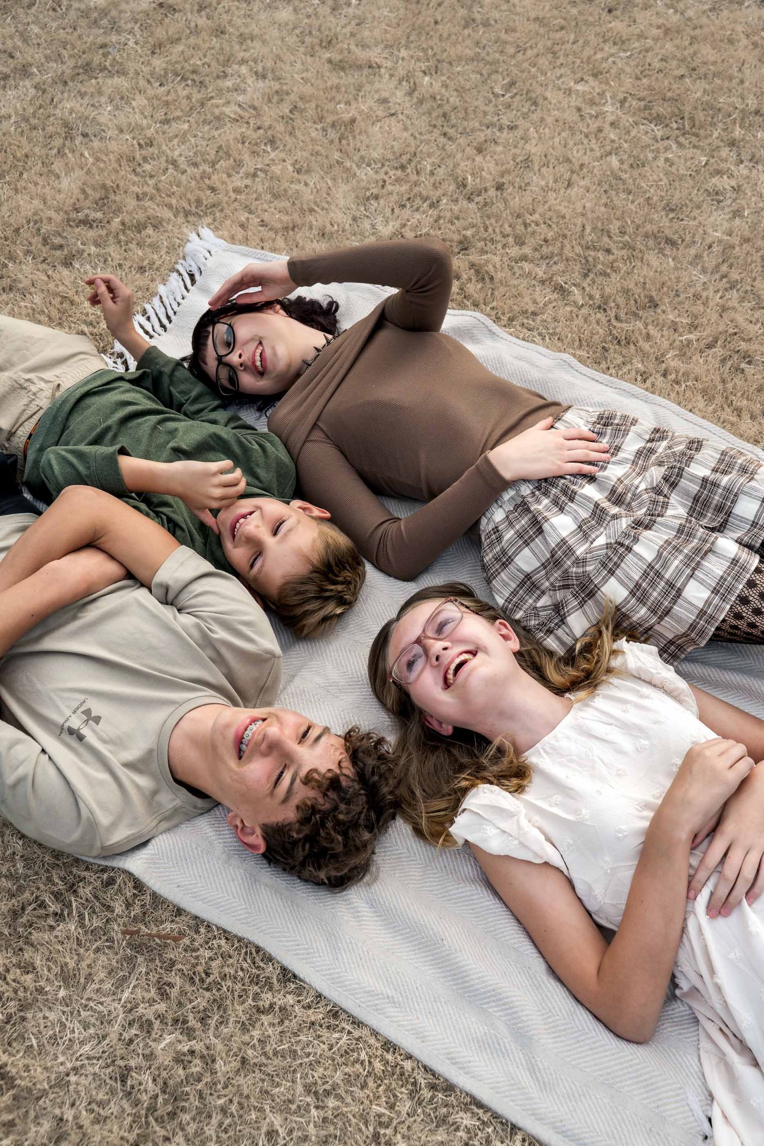 Four siblings laugh and smile while lying together on a white blanket on dry grass during outdoor family photos.