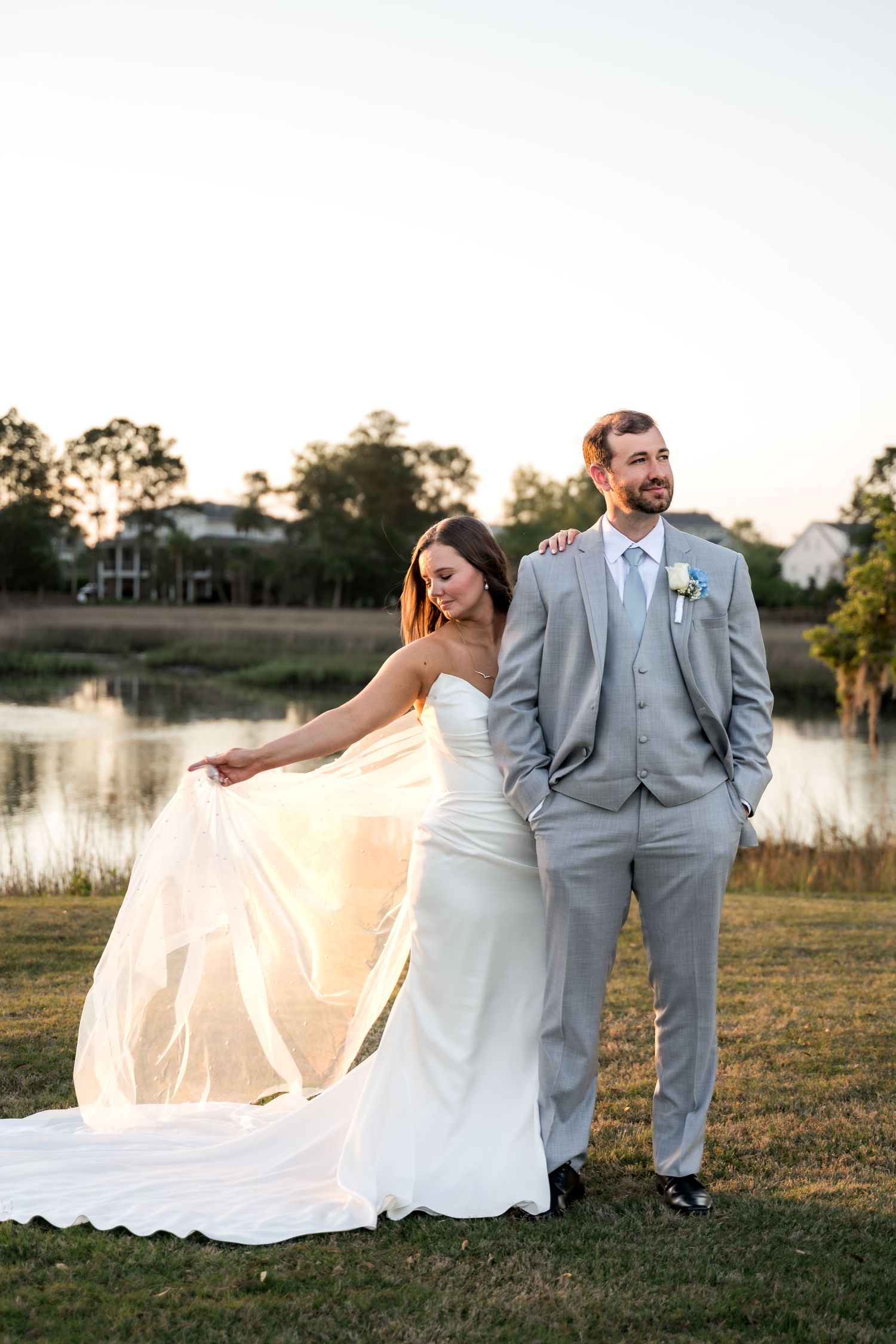 Bride twirls her veil while groom stands beside her near a lake at sunset in gray suit.
