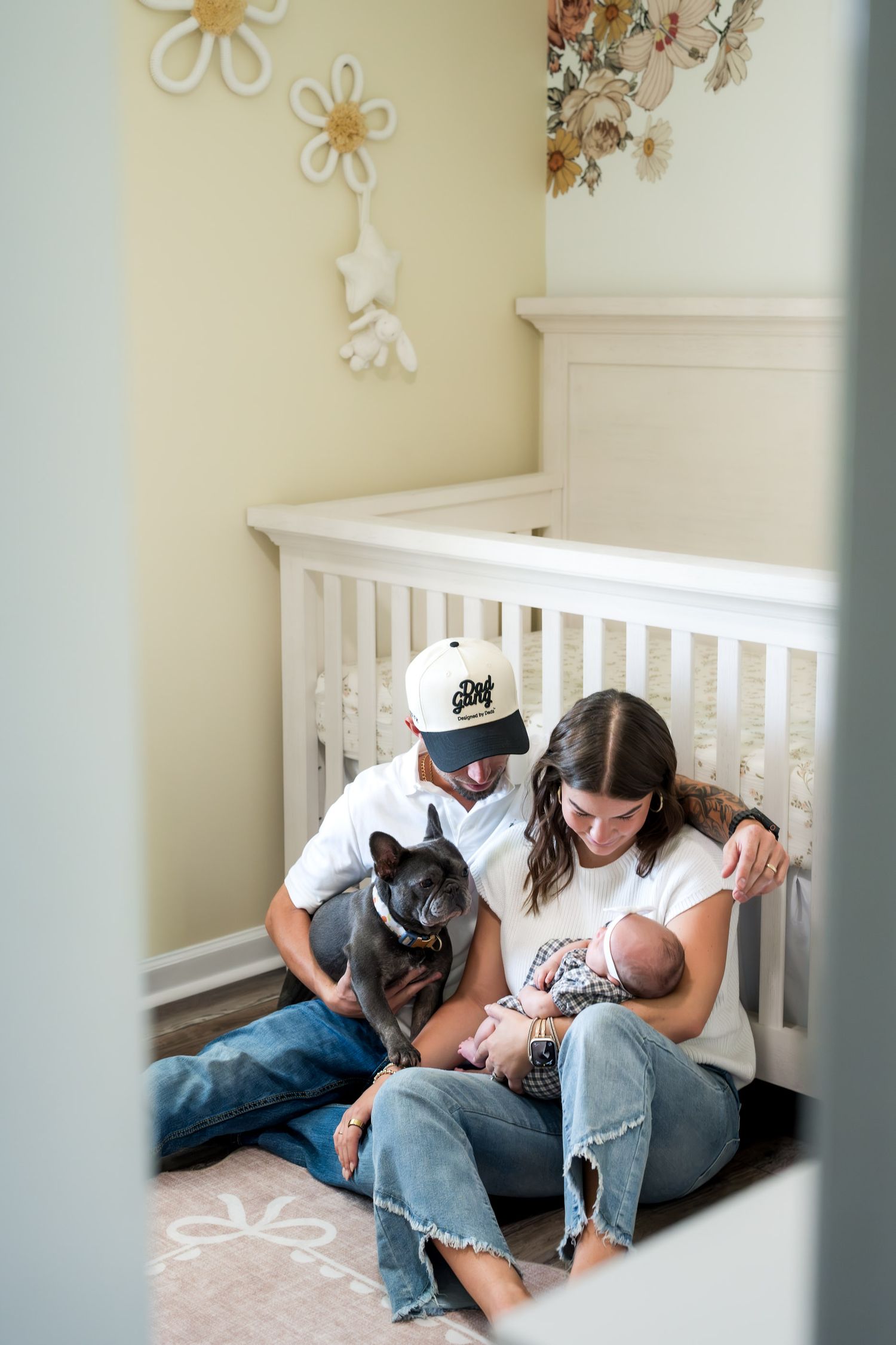 A couple sits on the nursery floor with their French Bulldog, lovingly gazing at their newborn baby by a white crib.