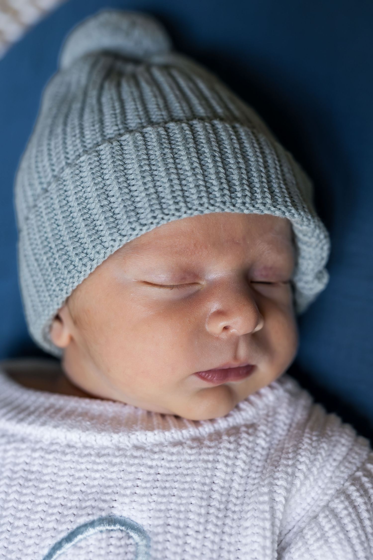 Peaceful newborn baby sleeping in a soft white outfit and light blue knit pom-pom hat.