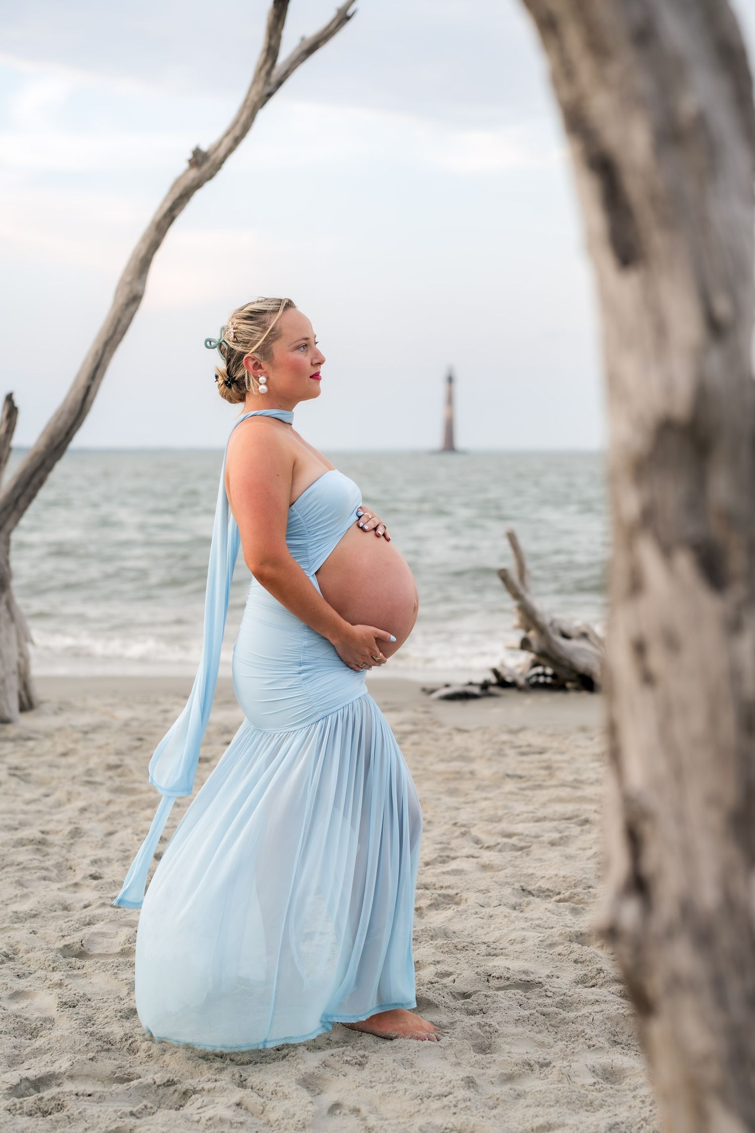 Pregnant woman in light blue gown on beach near lighthouse, cradling baby bump during maternity photoshoot.