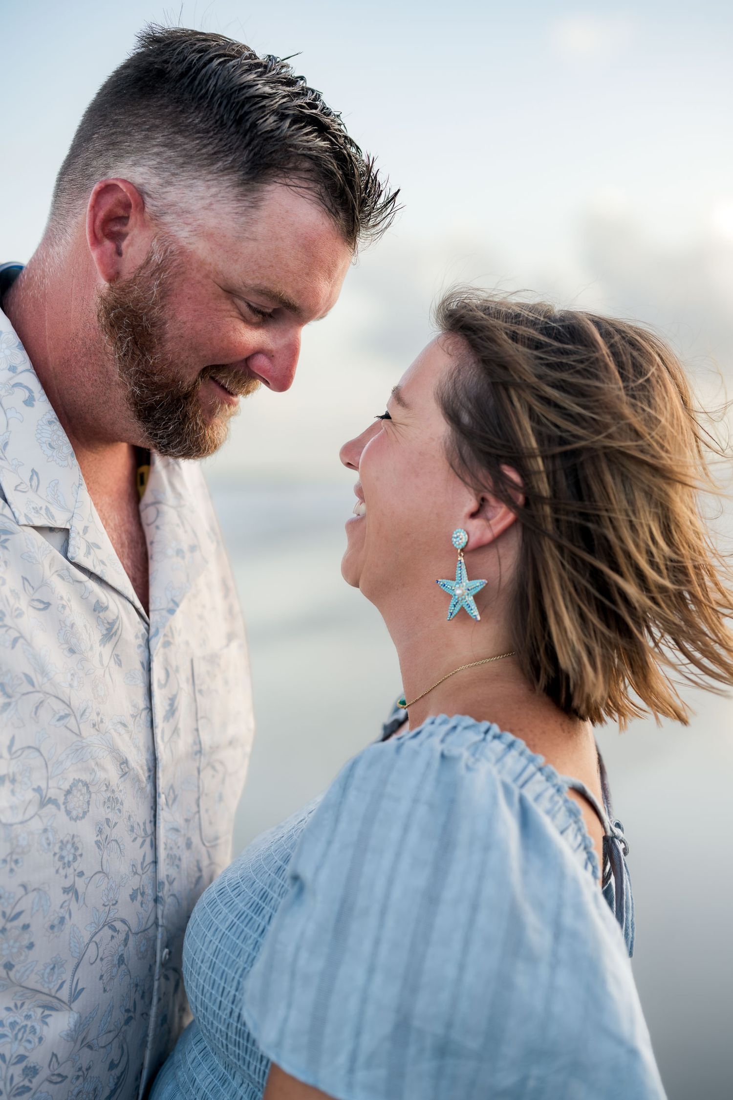 A couple shares a romantic moment on the beach, smiling at each other, woman wearing turquoise starfish earrings.