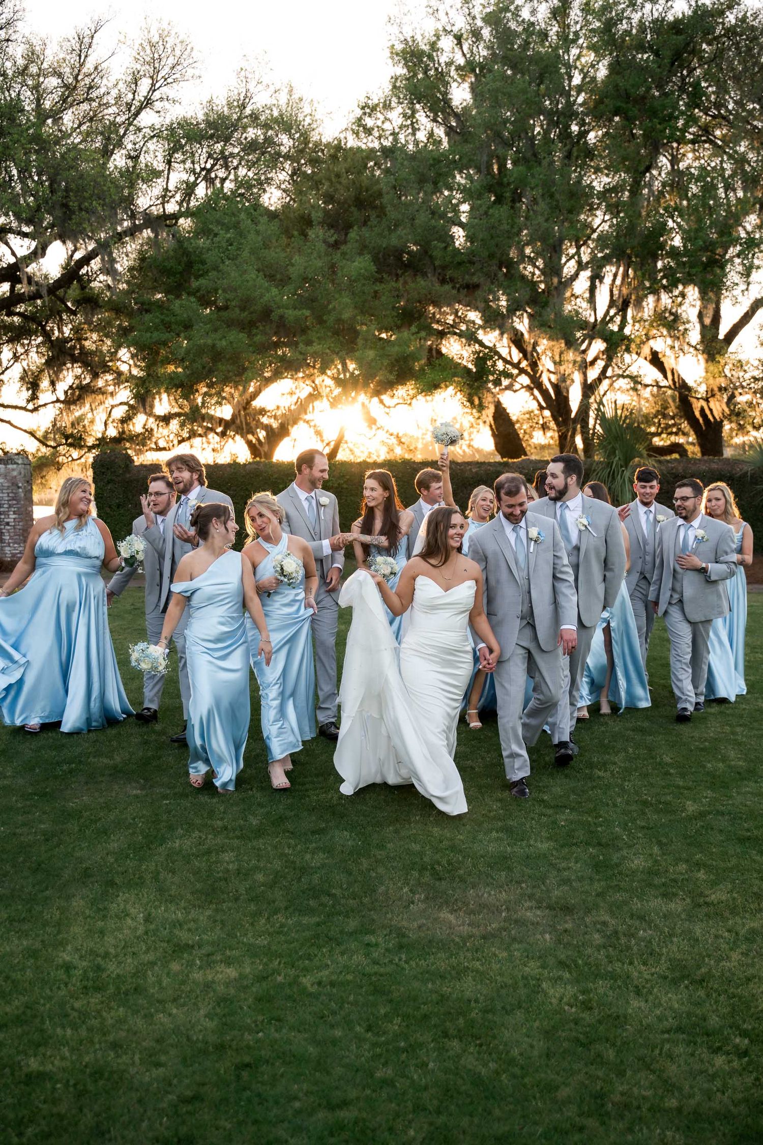 Dunes West. Wedding party walking together at golden sunset, bridesmaids in light blue satin gowns and groomsmen in gray suits.
