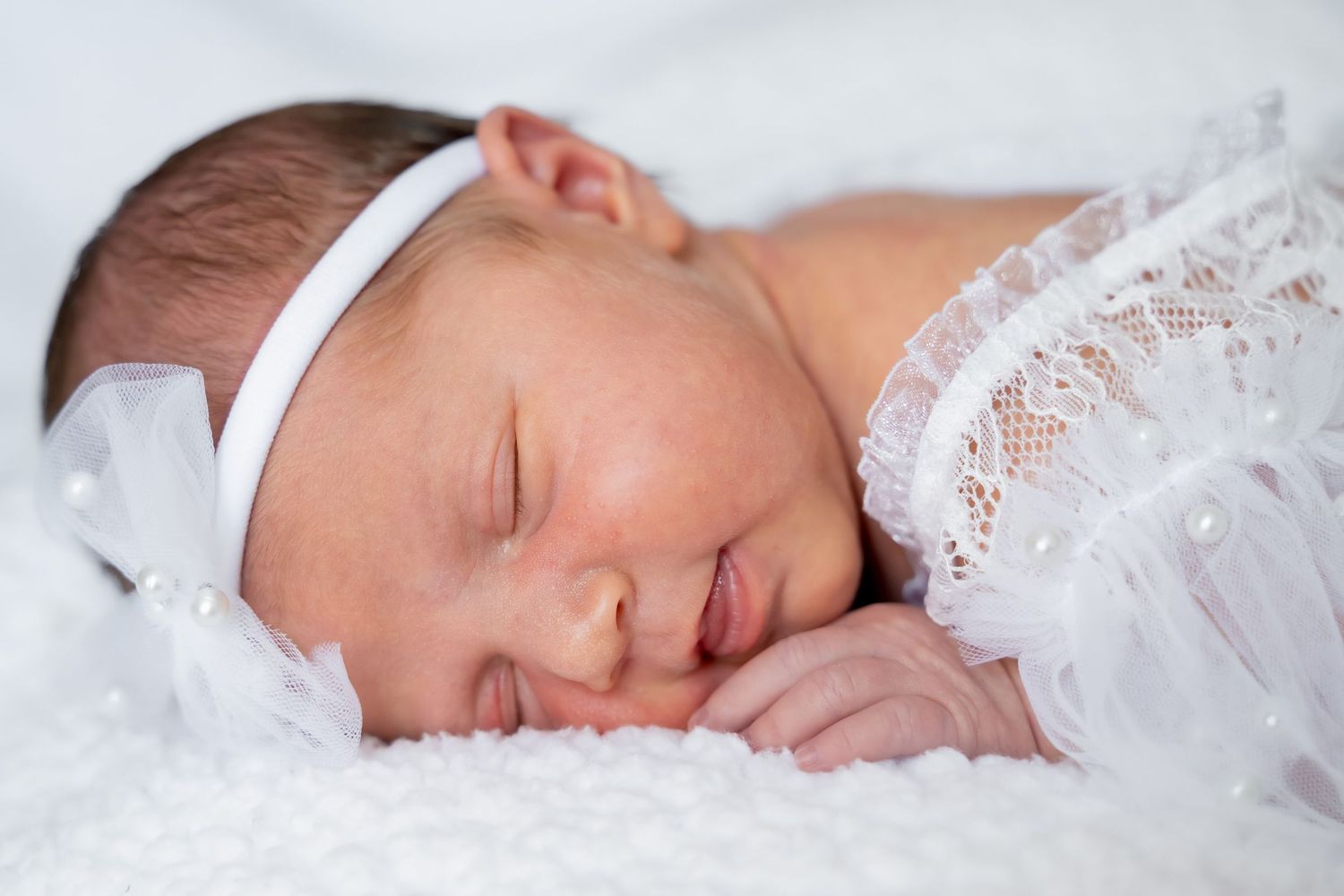 Sleeping newborn baby girl in white lace outfit and bow headband on soft white blanket.