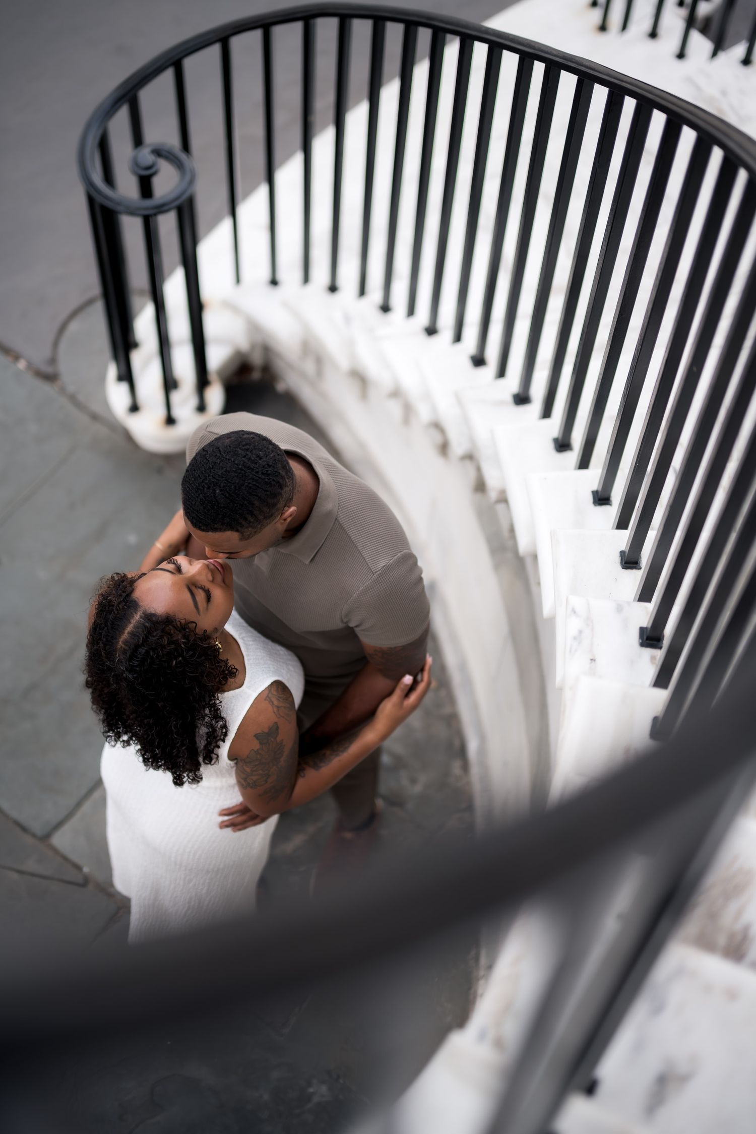 Aerial view of a couple sharing a kiss on elegant white spiral staircase with black iron railings.