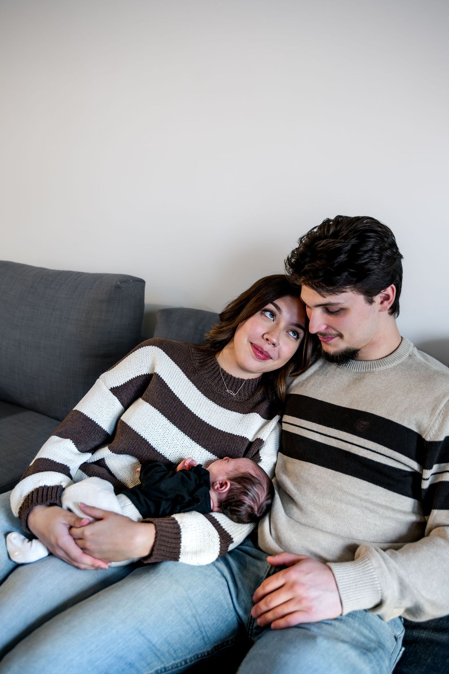 Young couple in striped sweaters sitting on gray sofa holding their sleeping newborn baby.