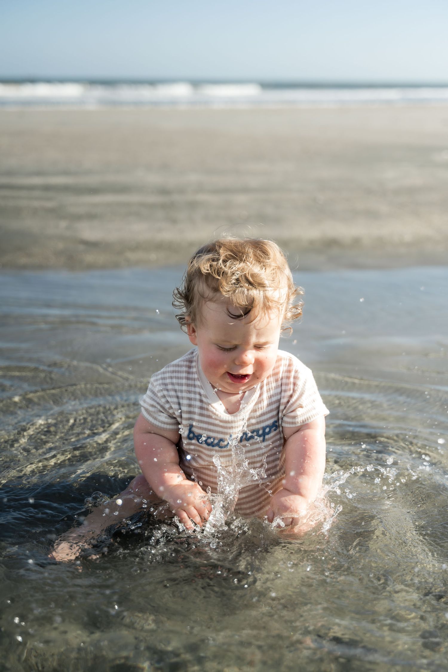 Isle of Palms. A happy baby in a striped onesie splashes and plays in shallow ocean water on a sunny beach.