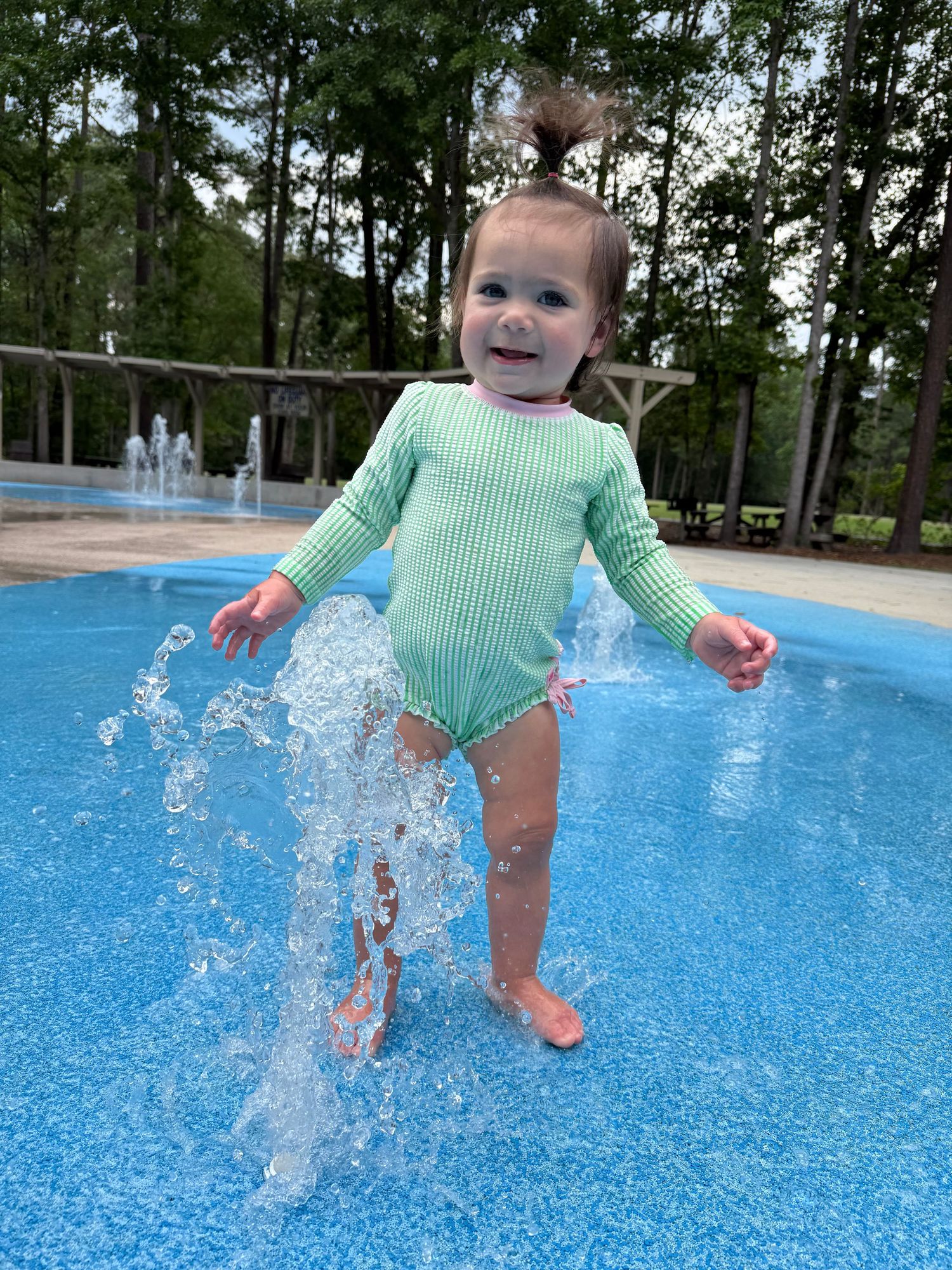 Smiling toddler girl in green striped swimsuit playing in splash pad water fountain outdoors.