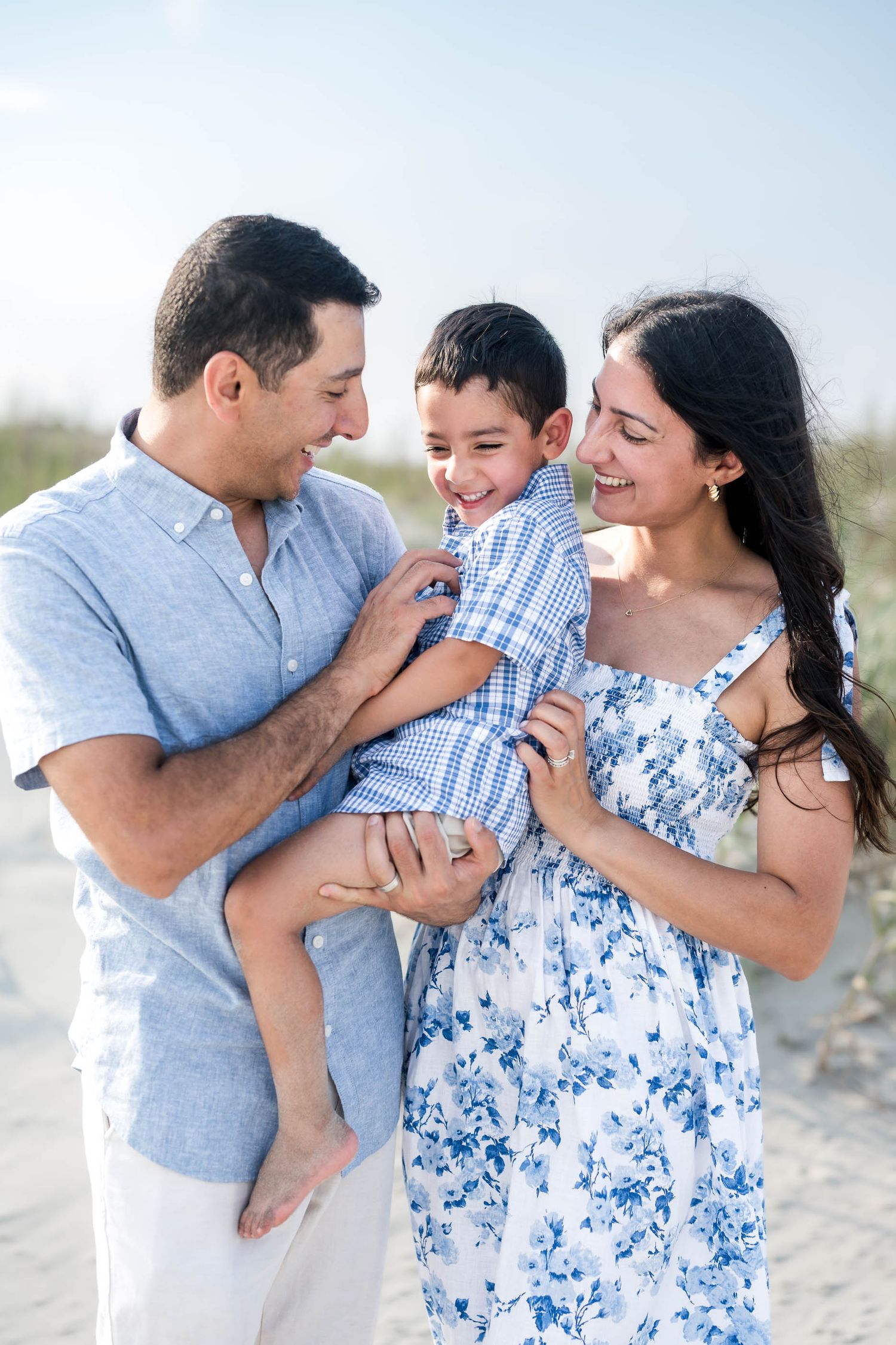Happy family of three sharing a joyful moment on a sunny beach, parents smiling at laughing boy in blue outfit.