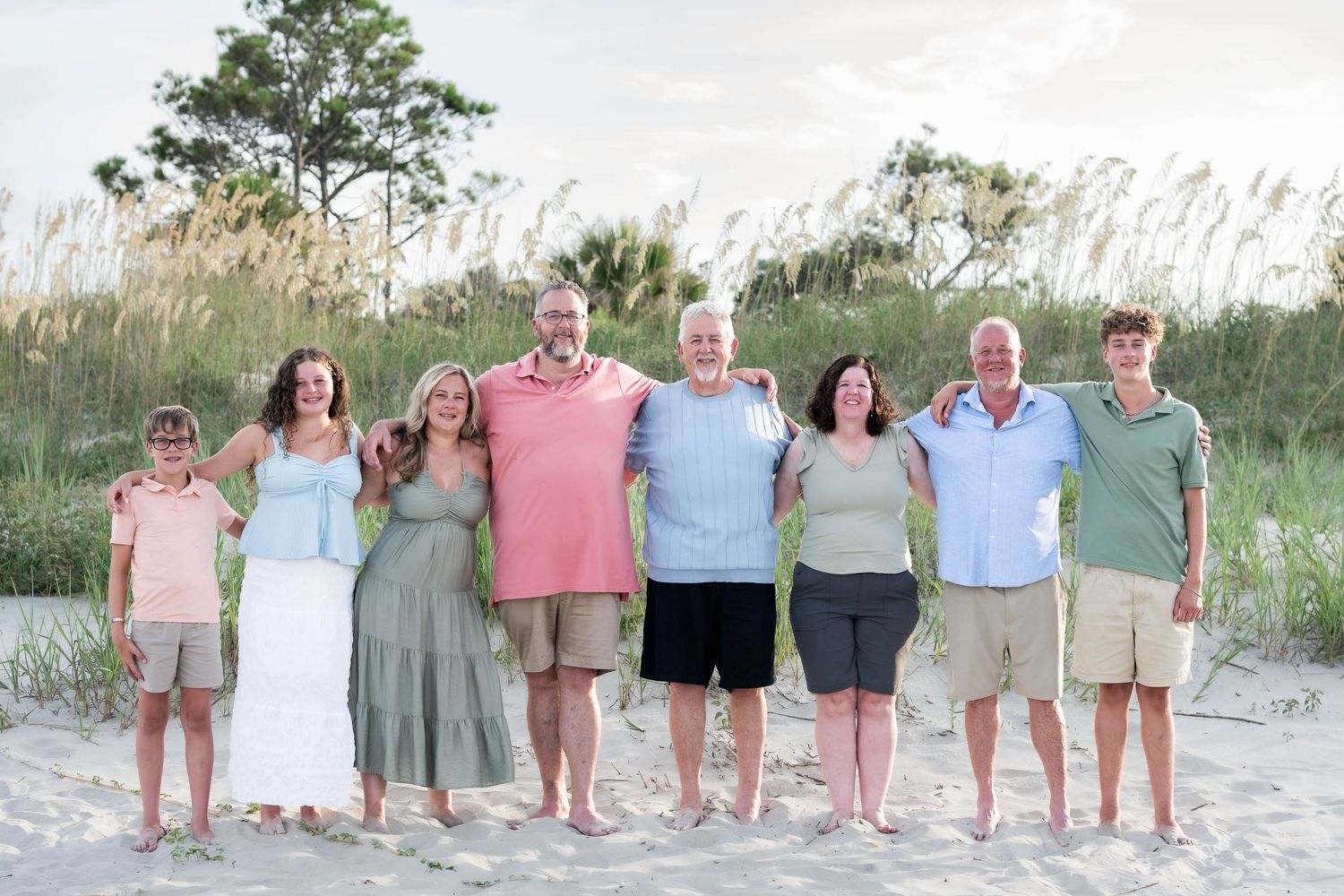 A multigenerational family of eight poses together on a sandy beach with sea oats in the background.