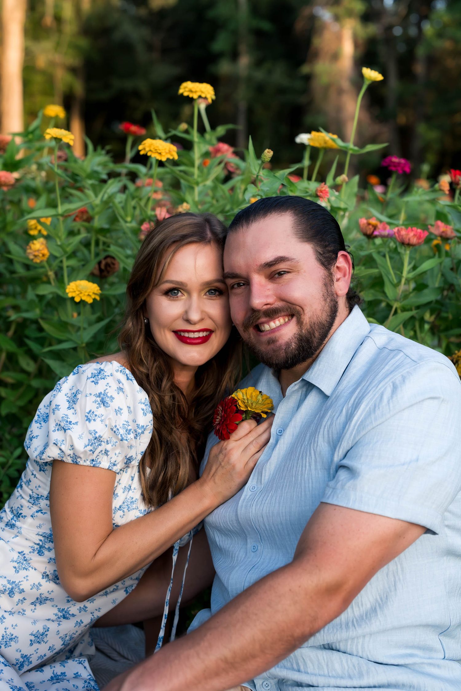 A smiling couple poses together in a colorful flower garden, the woman in a floral dress holding a red and yellow bloom.