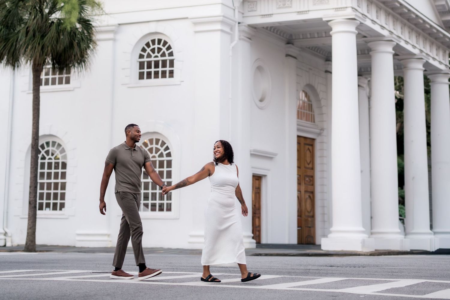 A couple holds hands walking in front of a white colonial building with tall columns on a sunny day.