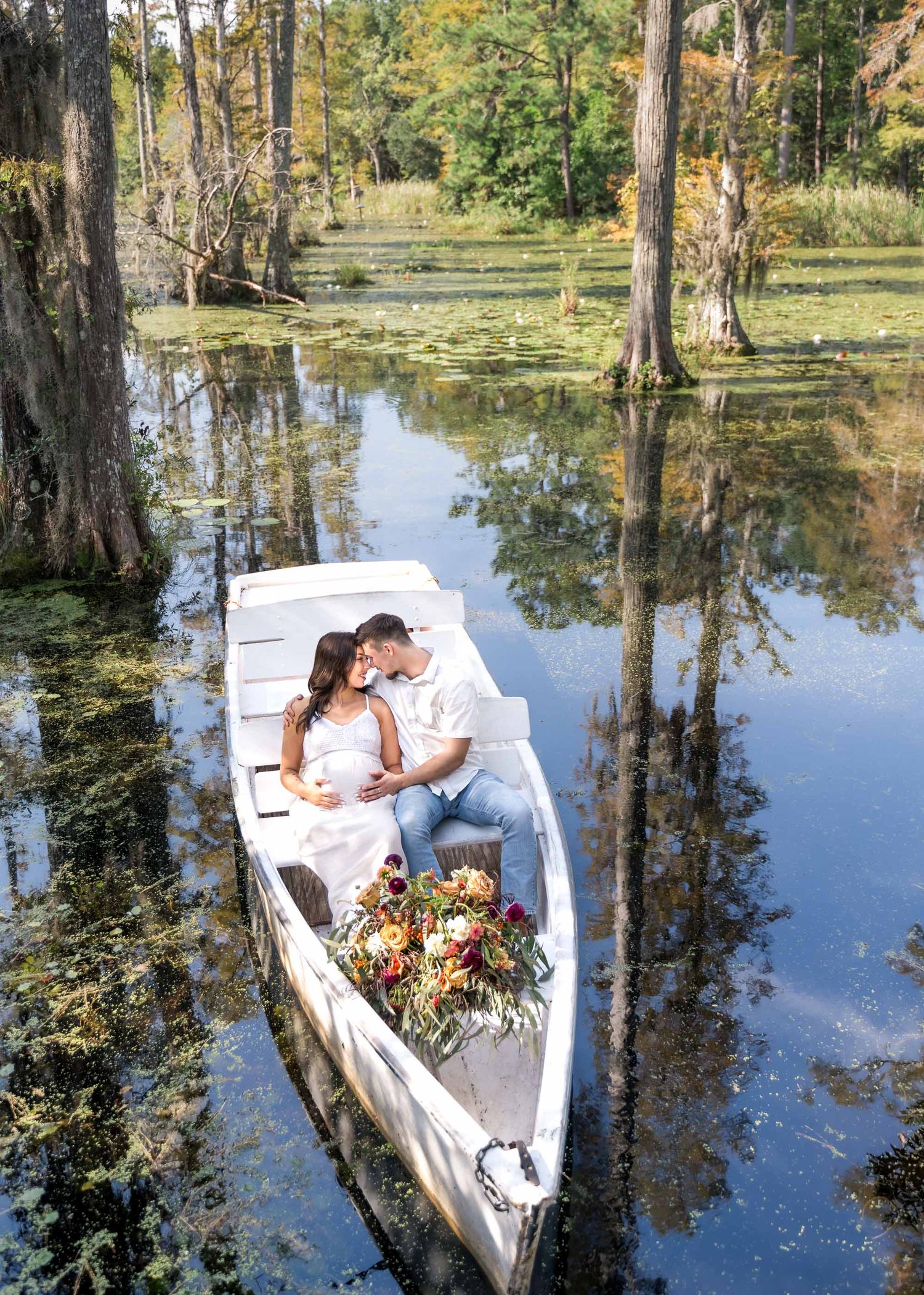 Couple sharing a romantic moment in a white rowboat on a serene cypress swamp, surrounded by lush greenery and a floral bouquet.