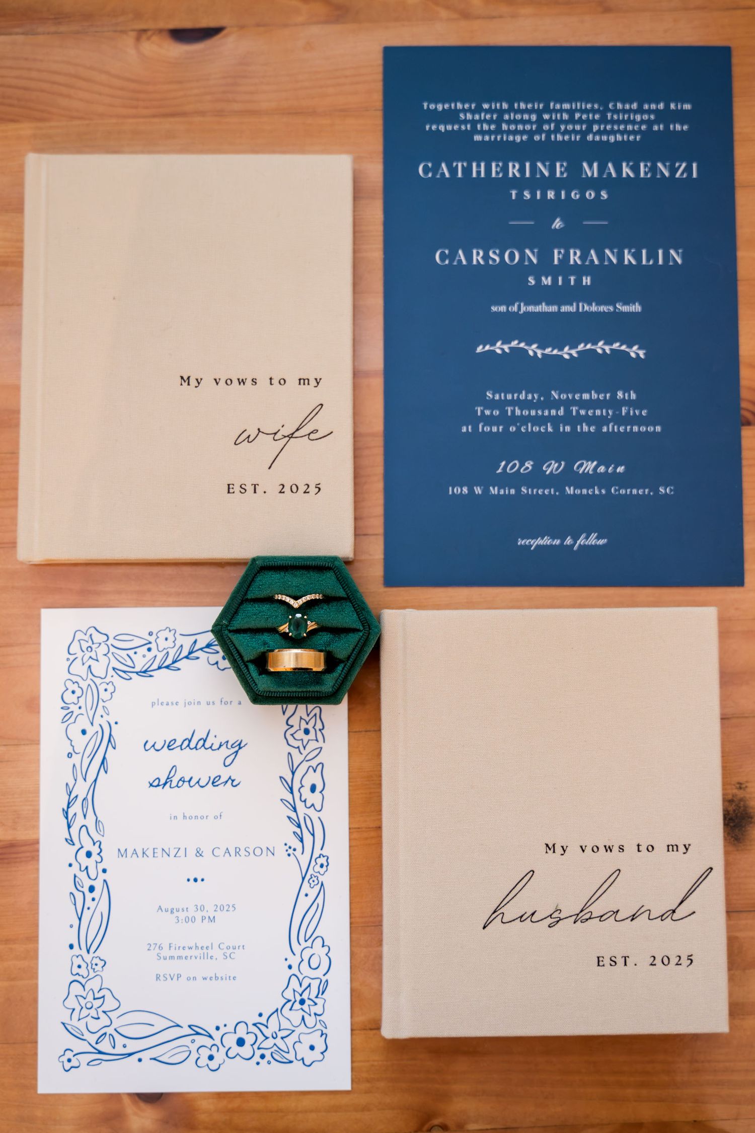 Elegant wedding stationery flat lay featuring navy blue invitations, vow books, and a green velvet ring box on wood.