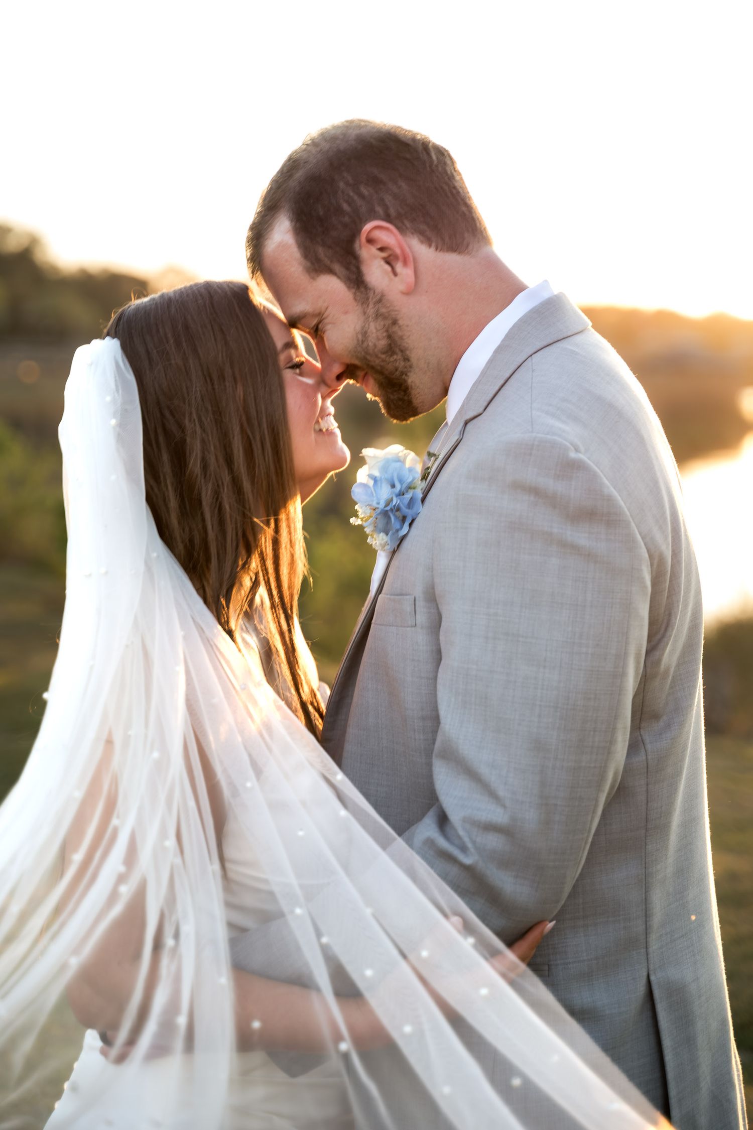 Dunes West Golf and River Club. Bride and groom sharing intimate forehead touch at golden sunset, bride in pearl veil, groom in gray suit.