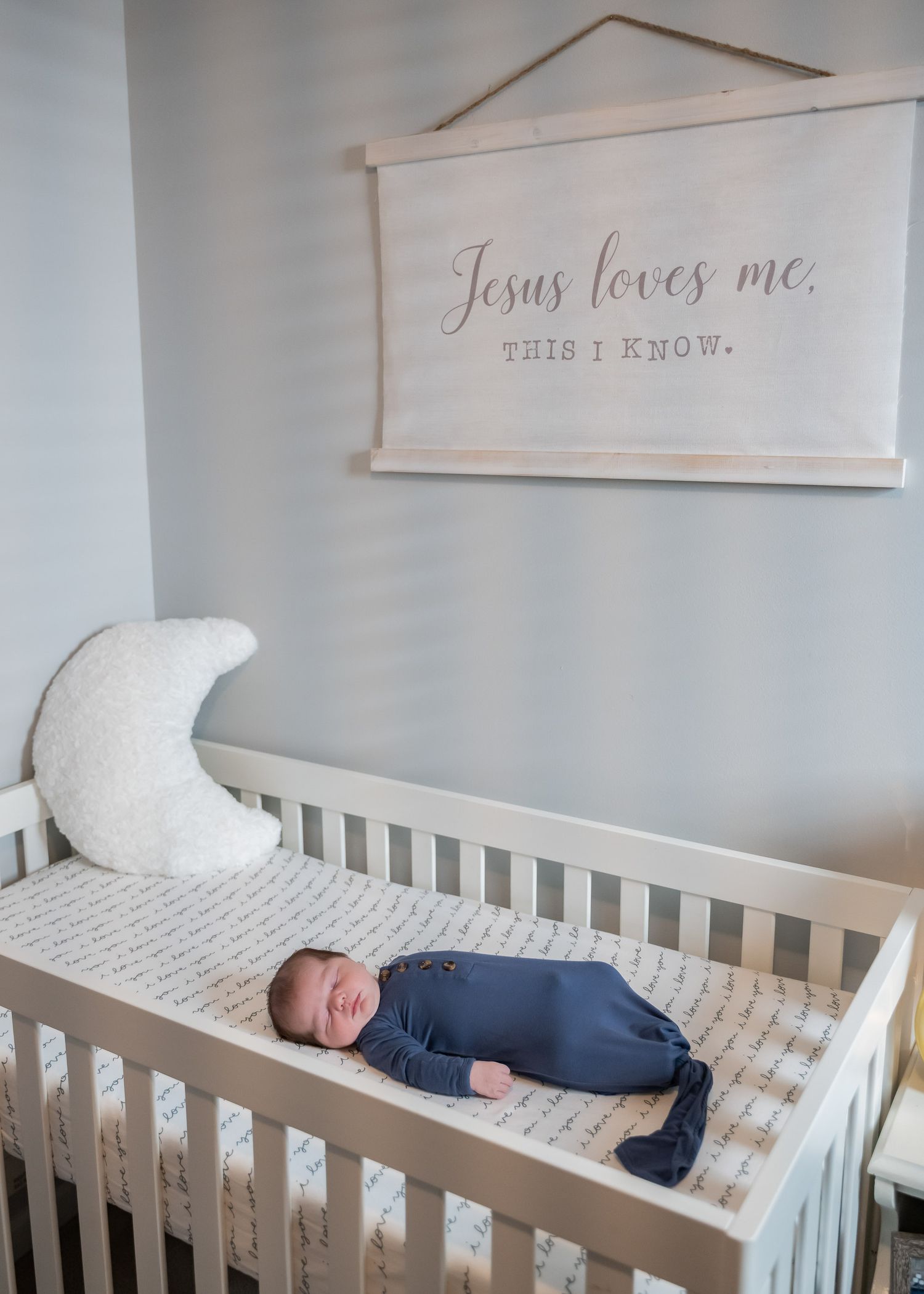 Newborn baby in blue wrap sleeping in white crib beneath Jesus Loves Me wall sign.