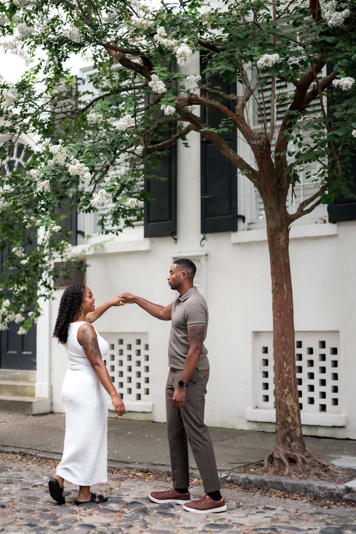 Couple holding hands and dancing beneath a blooming white tree outside a historic Charleston home.