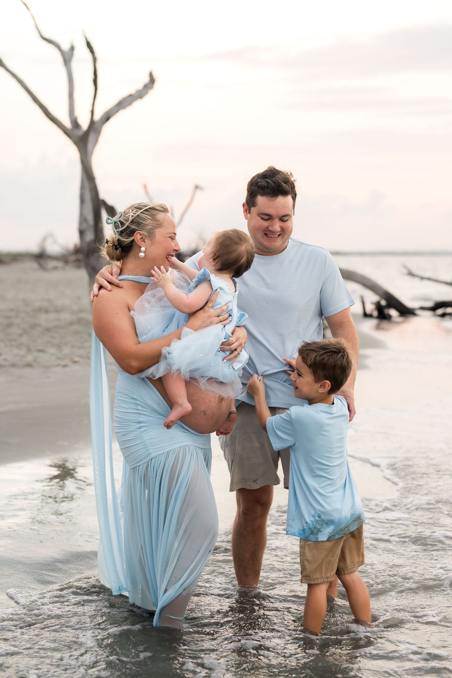 Family of four in light blue outfits wading in ocean water with driftwood backdrop during a beach photo session.