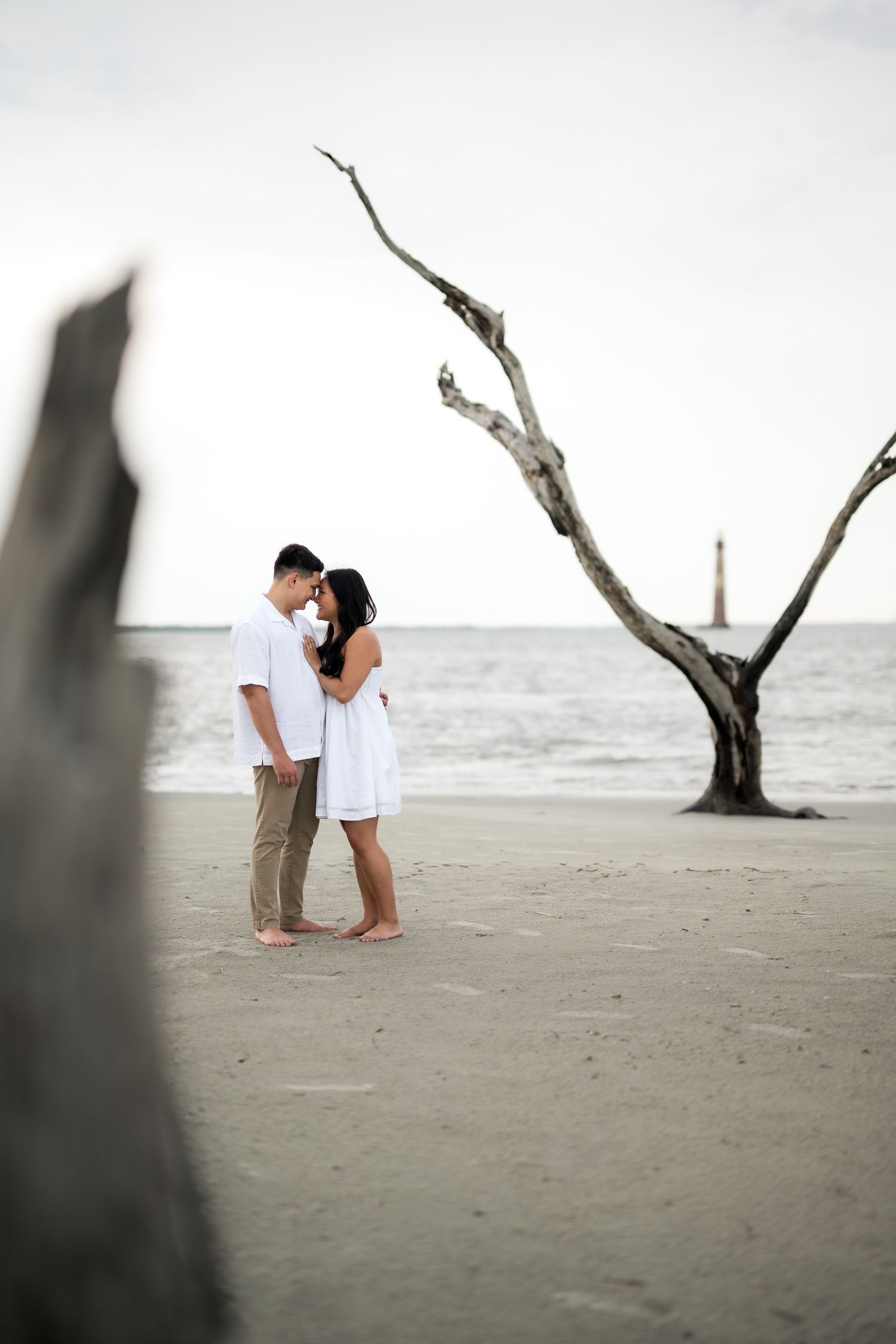 Couple embracing on a beach near a bare driftwood tree with a lighthouse in the background.