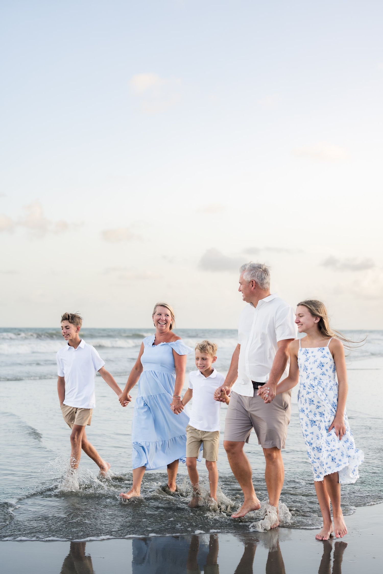 Multi-generational family holding hands walking barefoot on a beach shore at sunset in coordinated outfits.