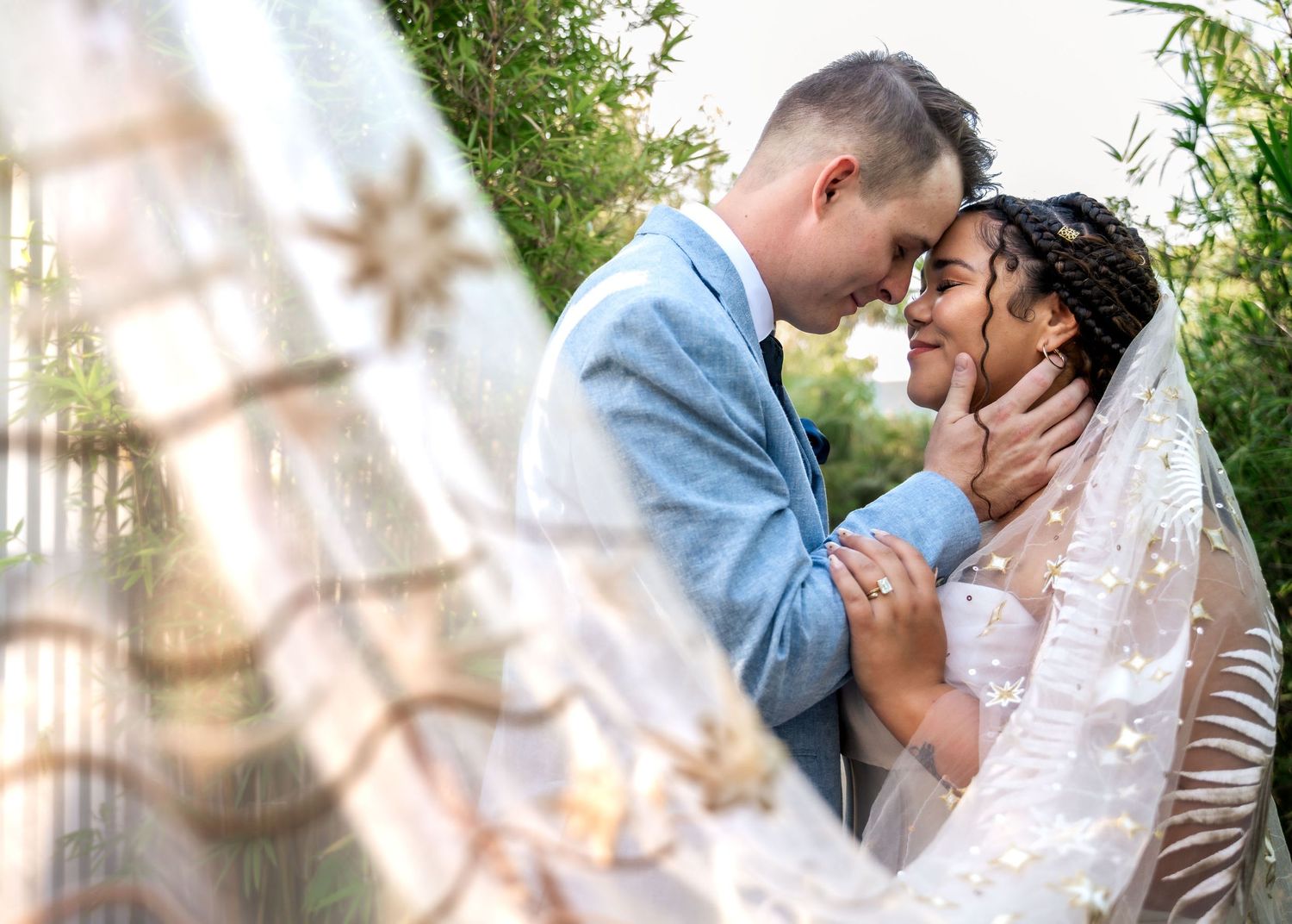 Romantic wedding couple sharing an intimate forehead touch moment surrounded by greenery beneath a decorative veil.