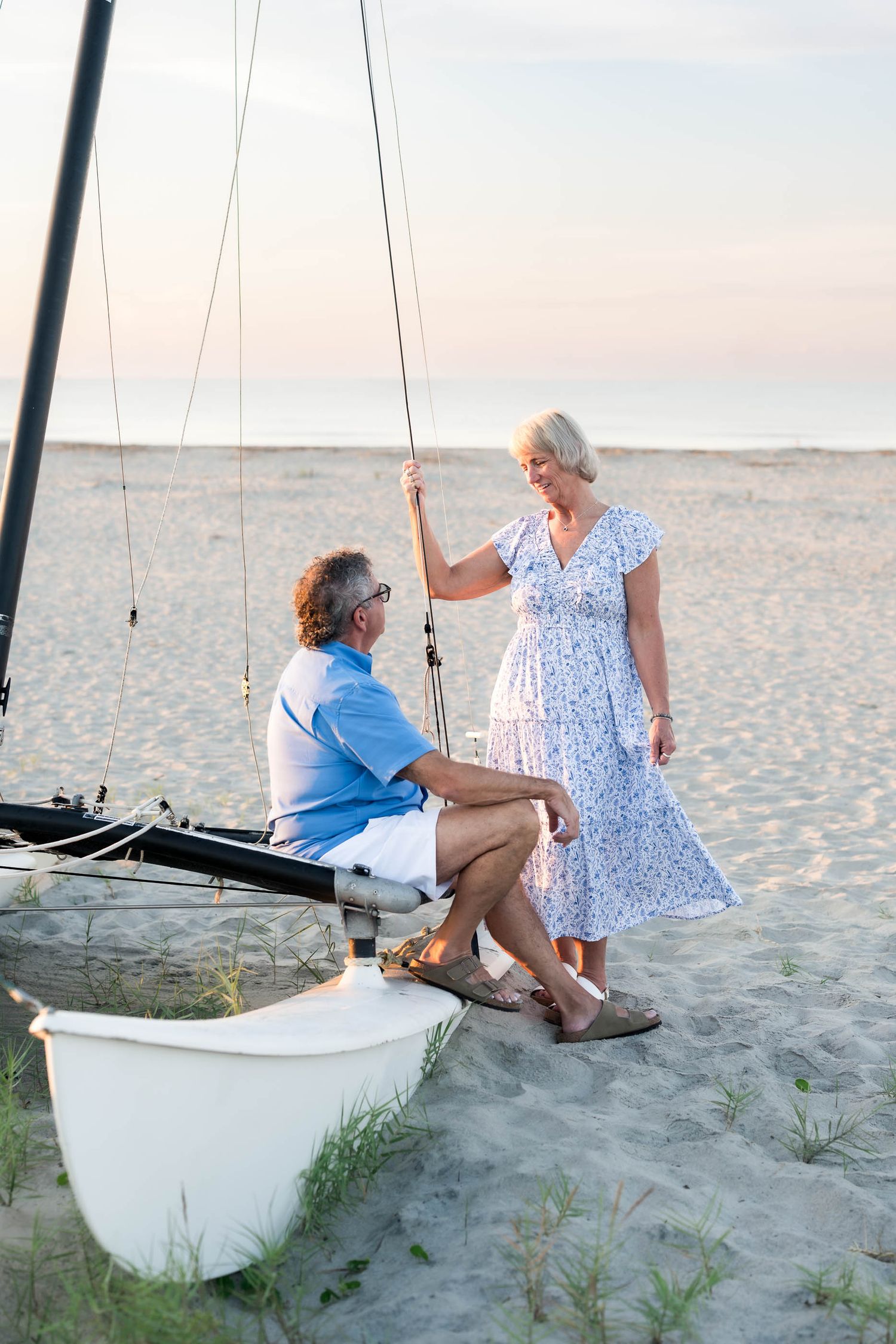 Older couple sharing a romantic moment by a sailboat on a sandy beach at sunset.