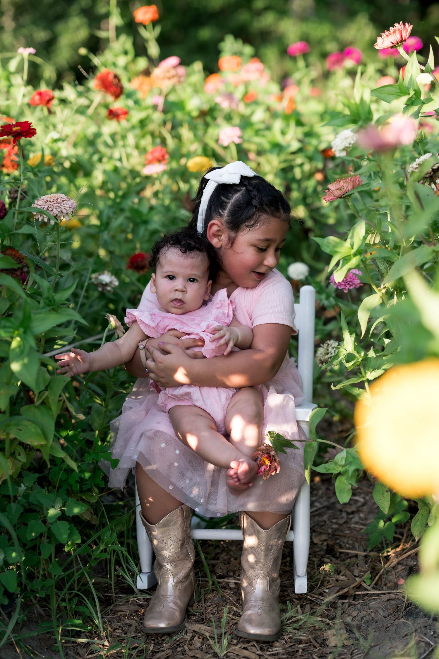 Two sisters in pink dresses sit in a white chair surrounded by colorful zinnia flowers in a garden.