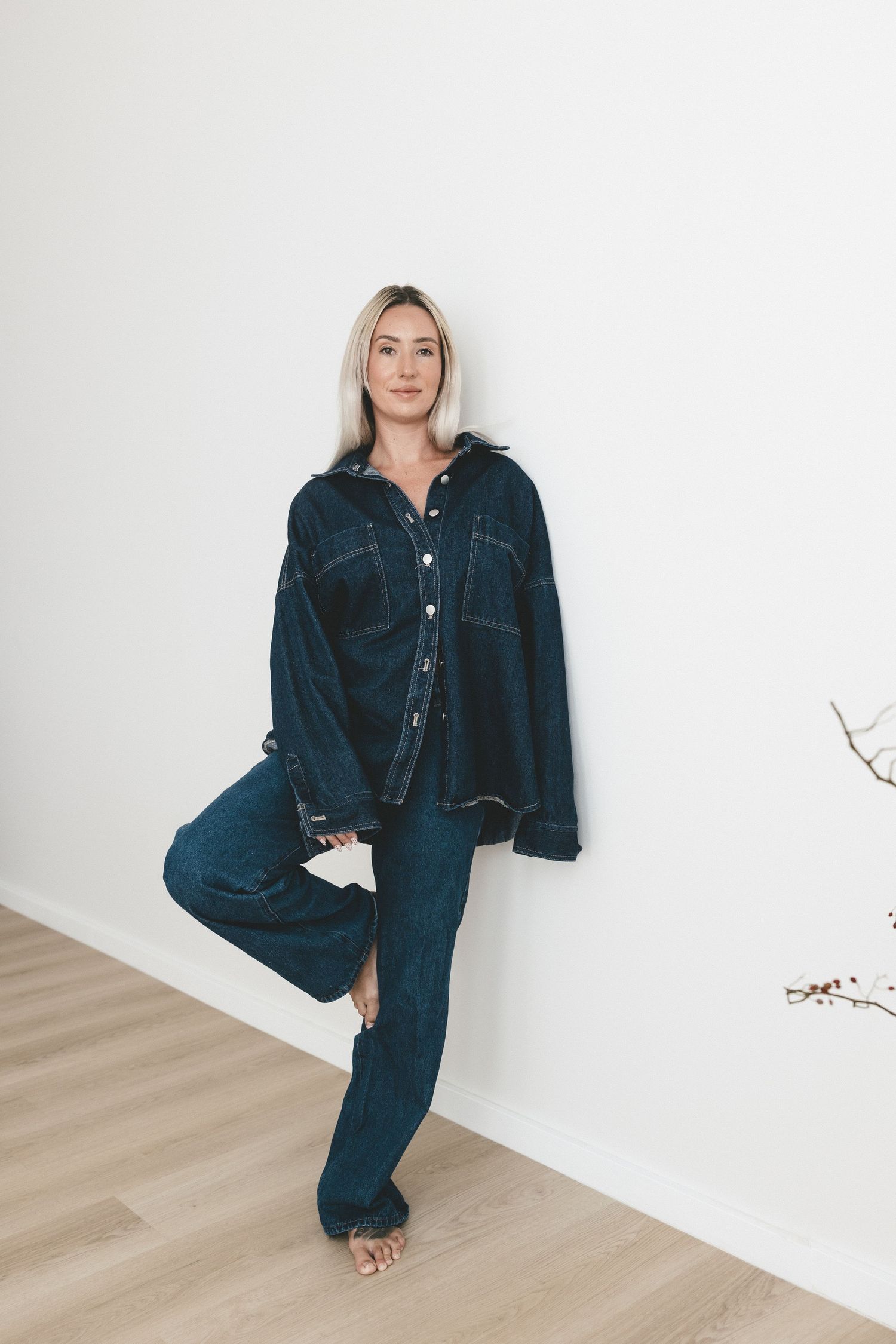 Woman in dark denim oversized jacket and wide-leg jeans posing against white wall on hardwood floor.