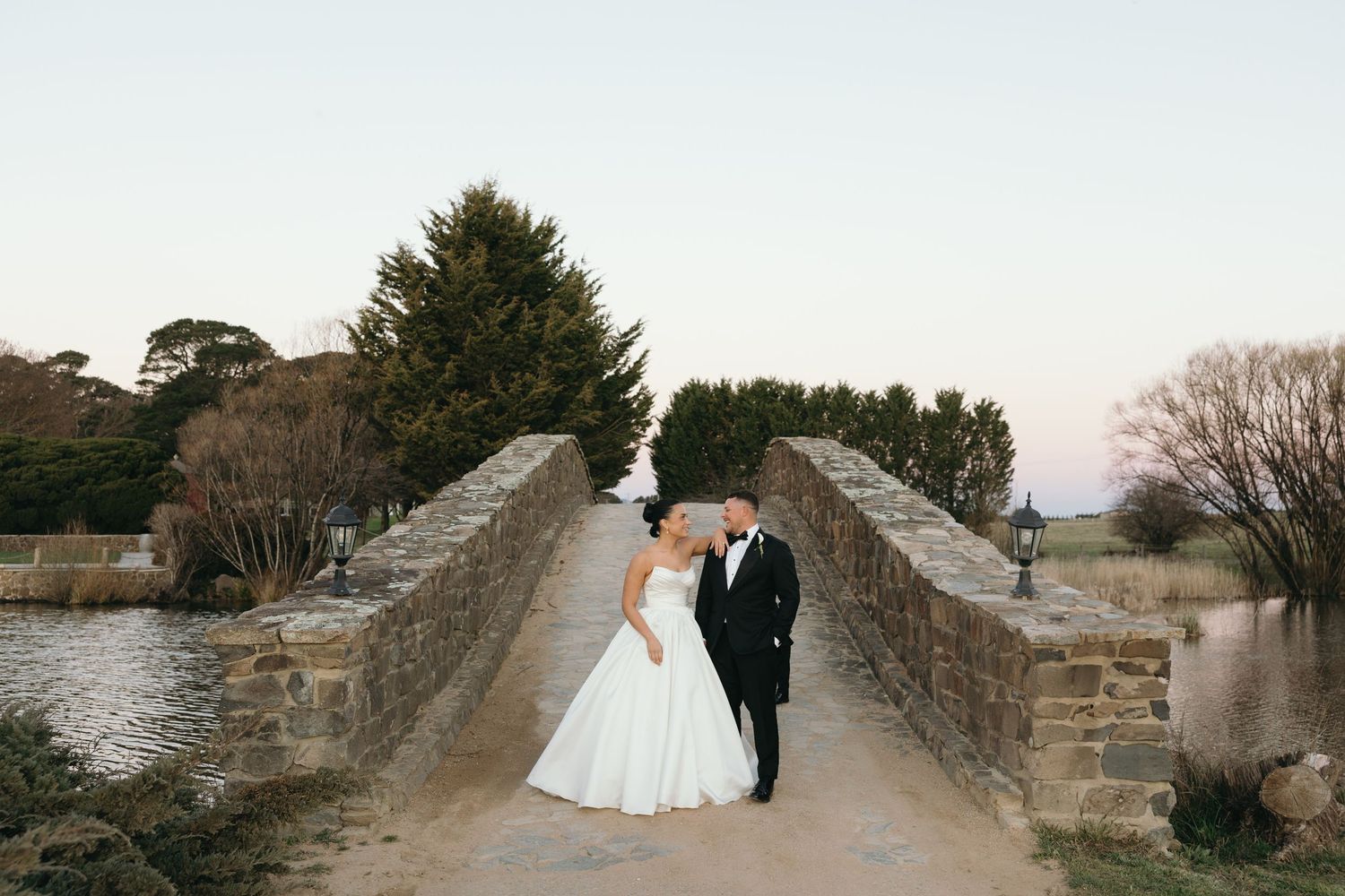 Bride and groom share a romantic kiss on a stone bridge at sunset during their outdoor wedding ceremony.