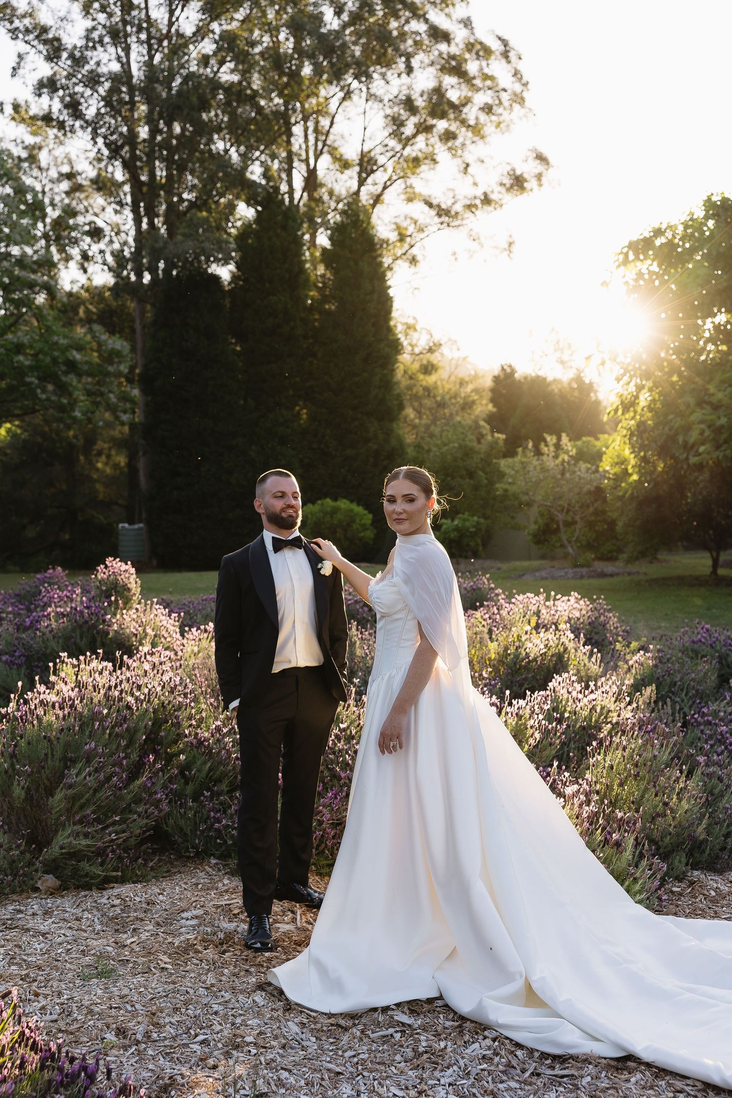Bride in white gown and groom in tuxedo pose in a sunlit garden with lavender blooms at golden hour.