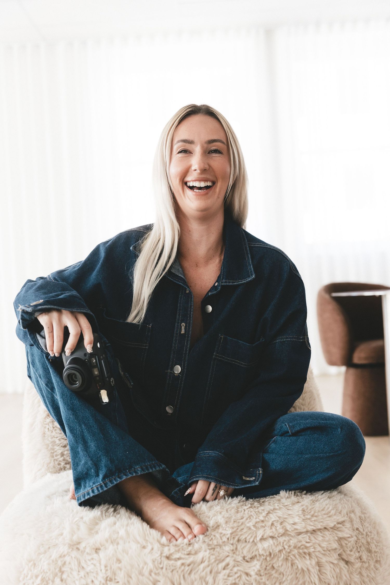 Smiling blonde woman in denim jacket and jeans sitting cross-legged on fluffy rug holding a camera indoors.