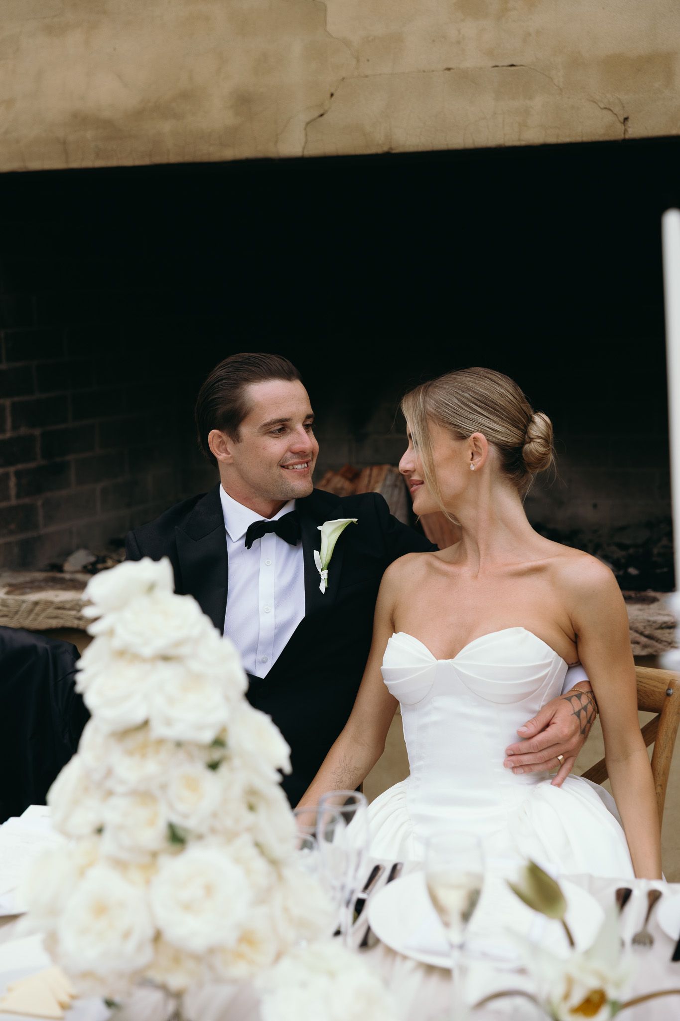 A smiling groom in a tuxedo and bride in a strapless white gown sit together at their wedding reception table.