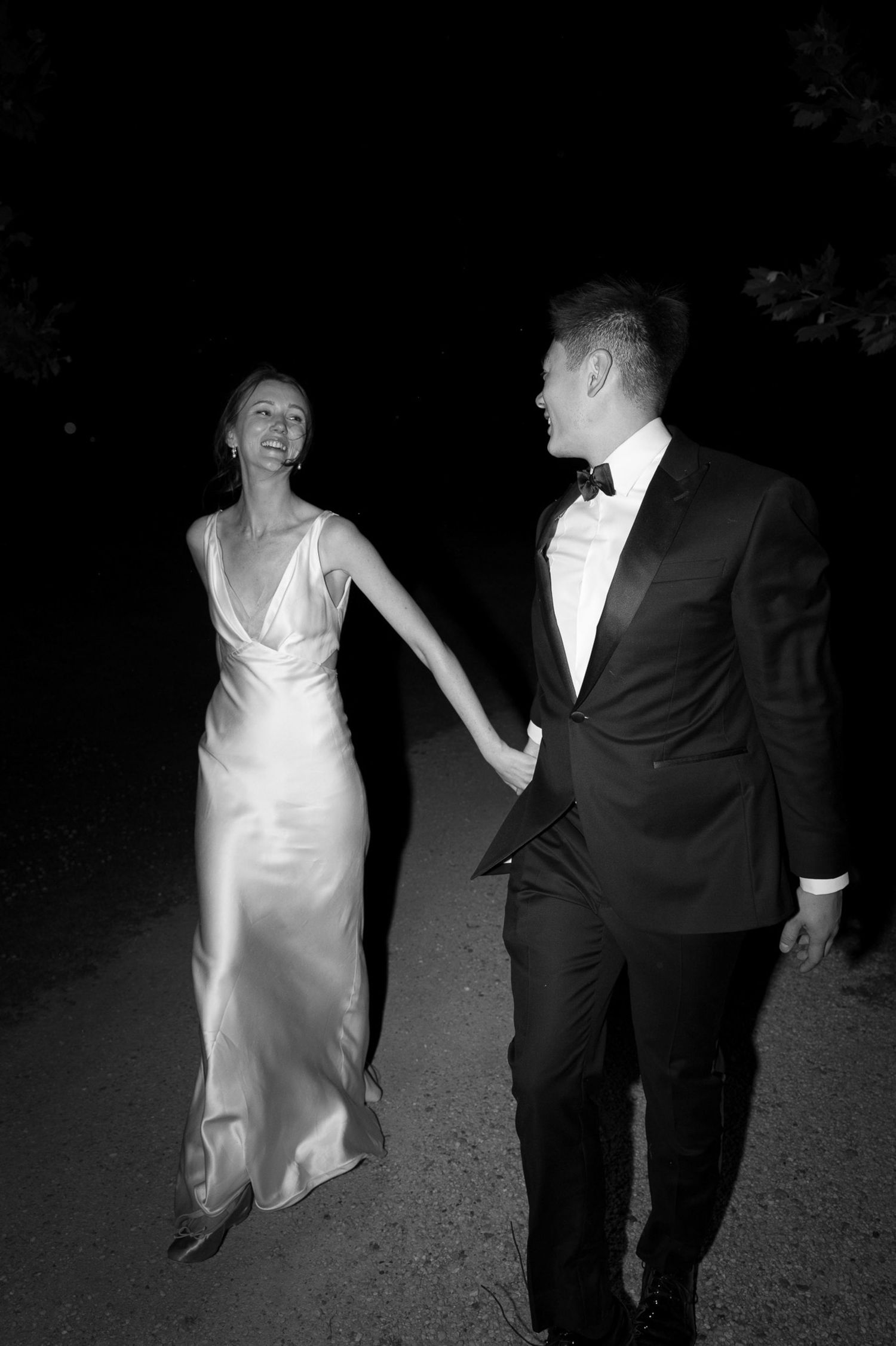 Black and white photo of a bride in satin gown and groom in tuxedo holding hands walking at night.