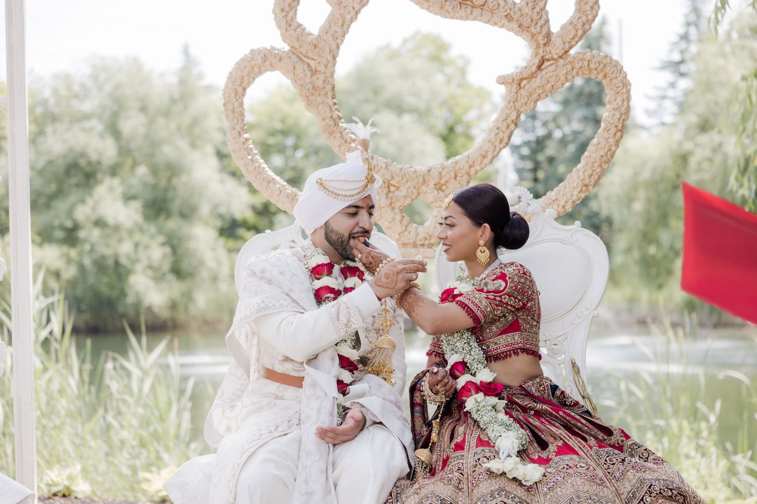 A couple in traditional wedding attire shares a romantic moment outdoors surrounded by lush greenery.
