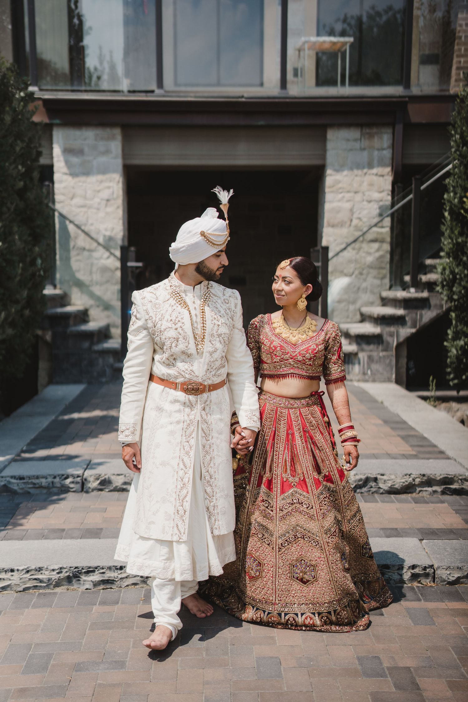 A couple in traditional South Asian wedding attire stand together on a stone walkway outside an elegant building.