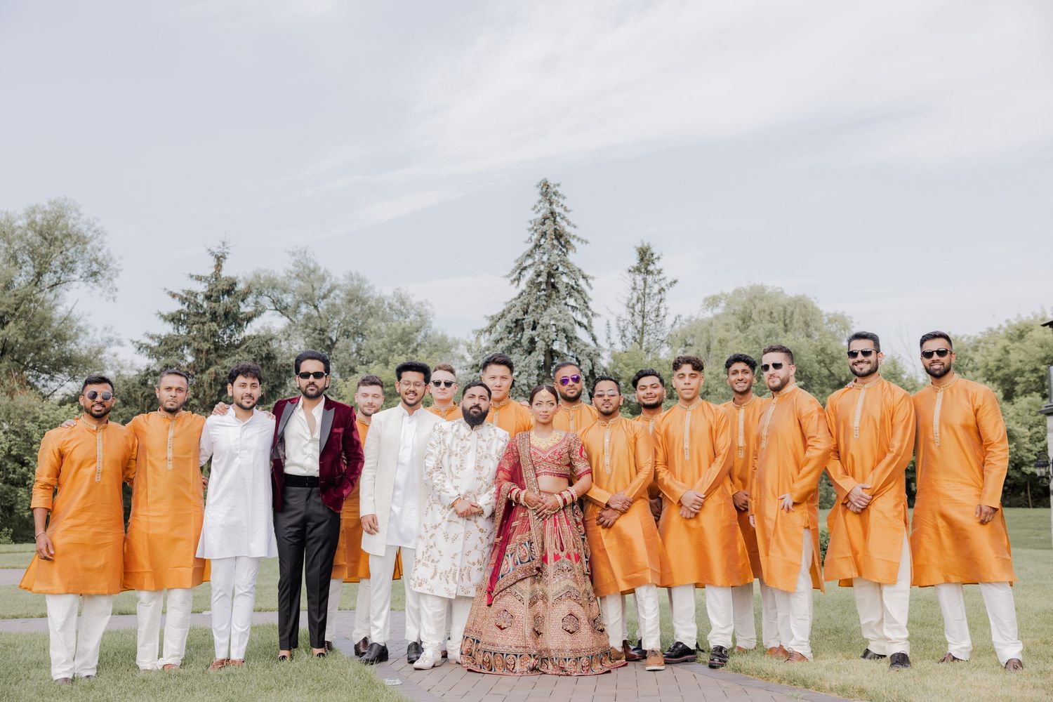 A wedding party poses outdoors in coordinated orange and white attire against a natural backdrop.