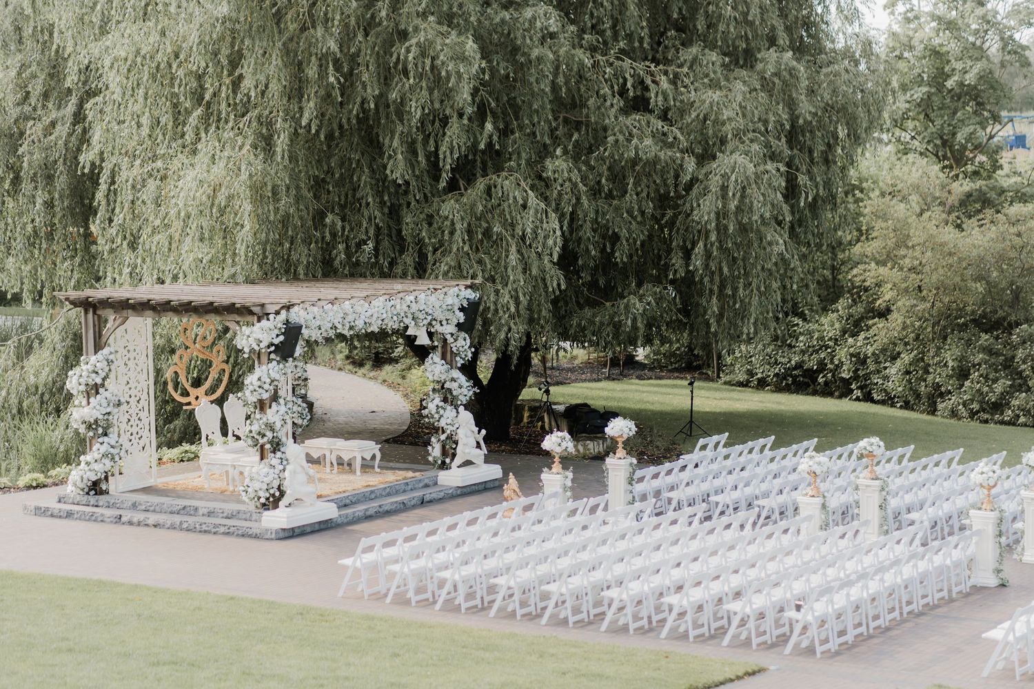 Outdoor wedding venue setup with rows of white chairs on a lawn surrounded by willow trees.