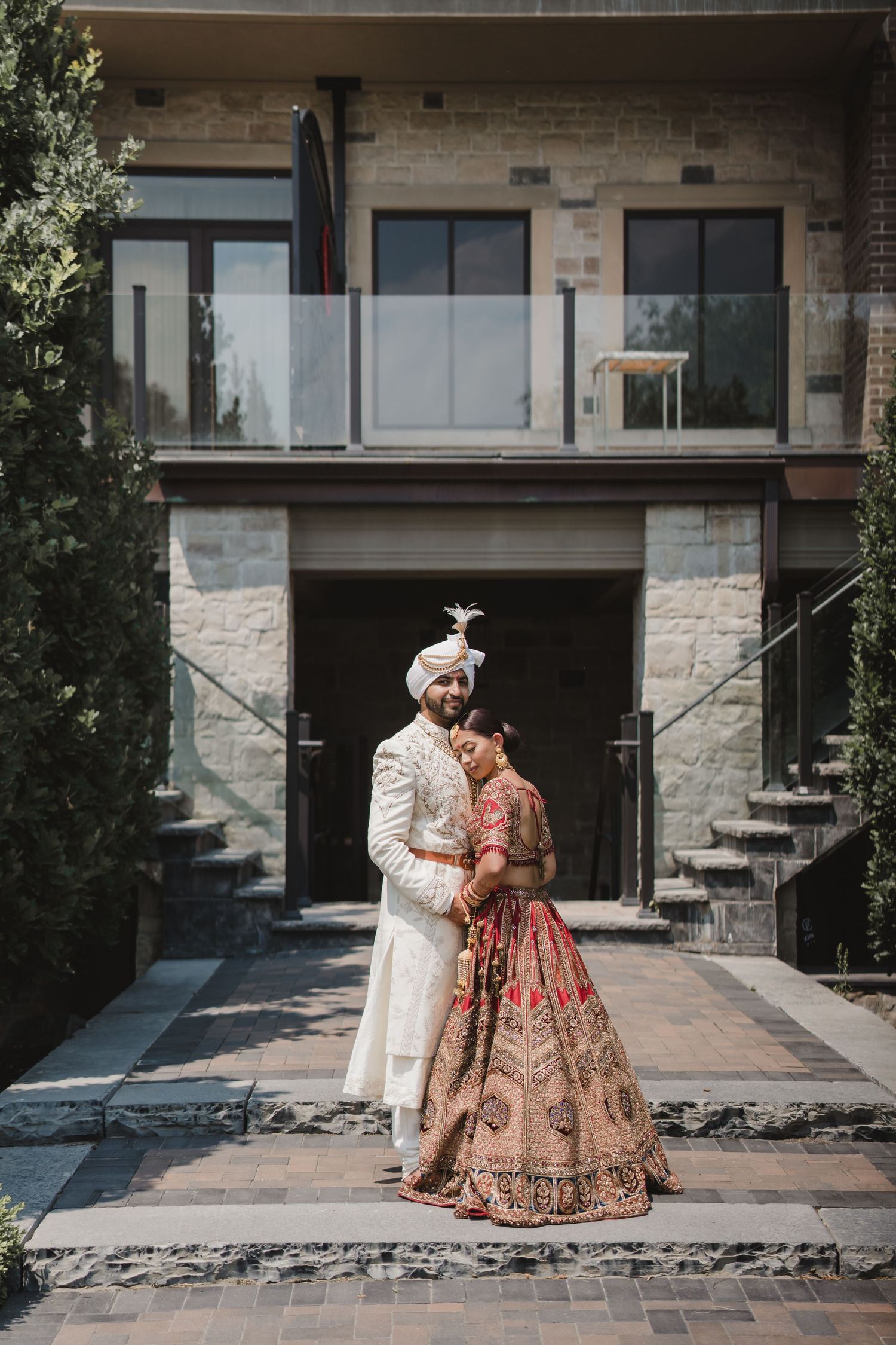 A couple in traditional South Asian wedding attire poses together against a modern stone building exterior.