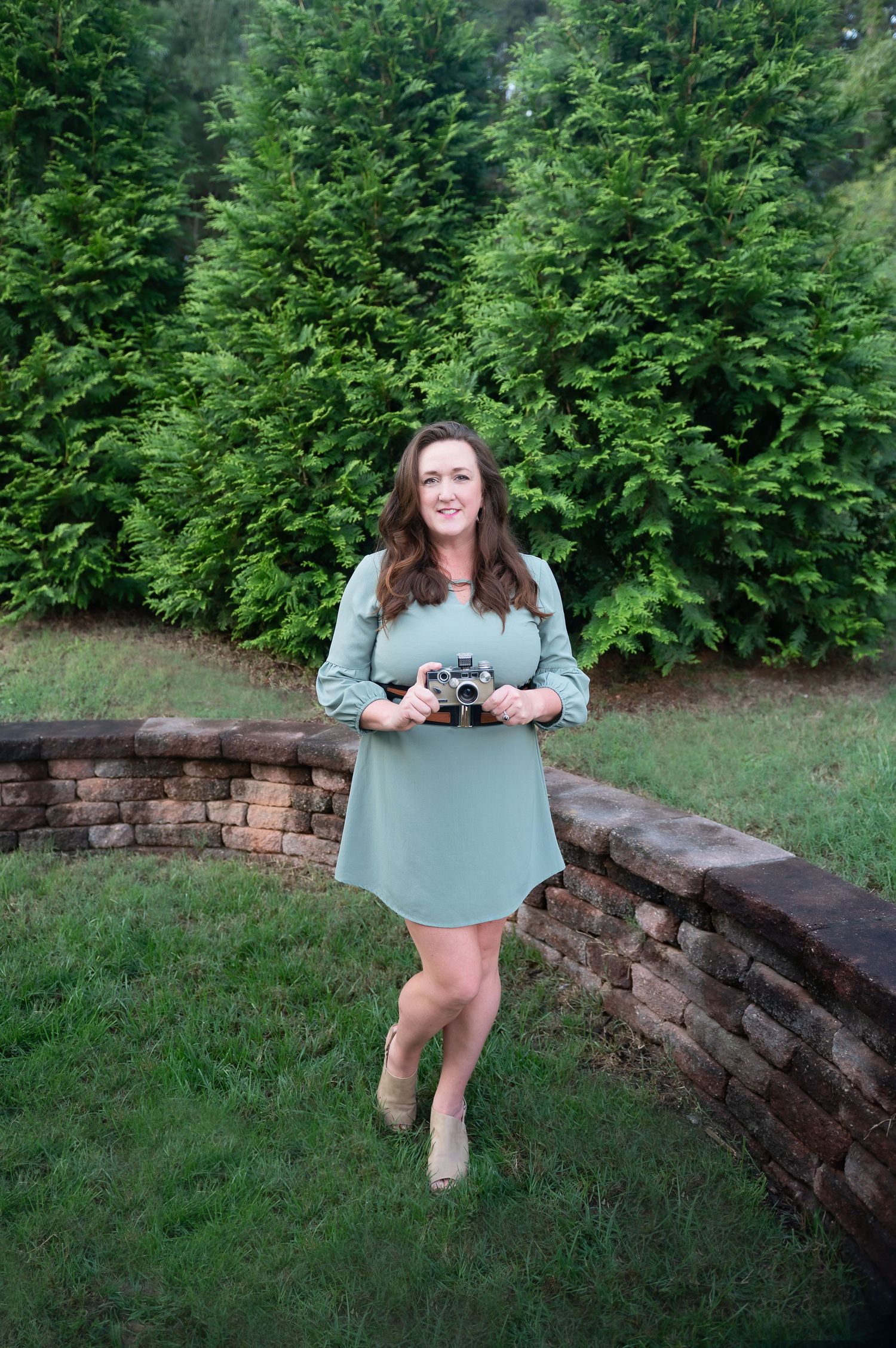 Person in mint green dress sitting on curved brick wall with evergreen trees in background.